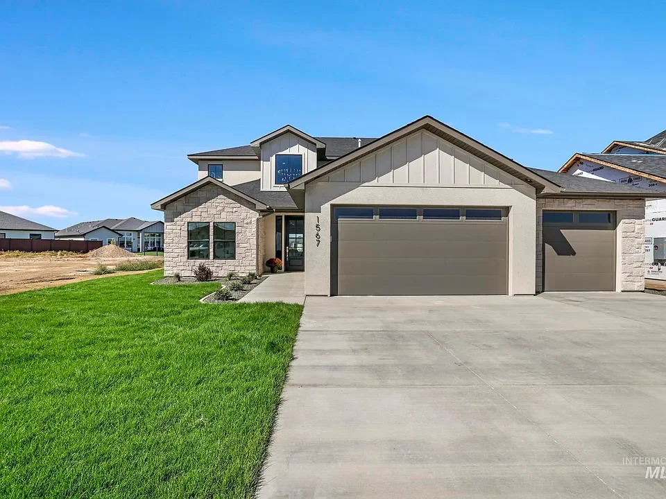 Modern two-story house with a large green lawn, concrete driveway, and attached garage. The house features a mix of stone and siding exterior with a blue sky background.