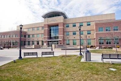 A large modern building with a curved glass structure on top, in front of a park with benches, lamp posts, and a trash can, under a partly cloudy sky.
