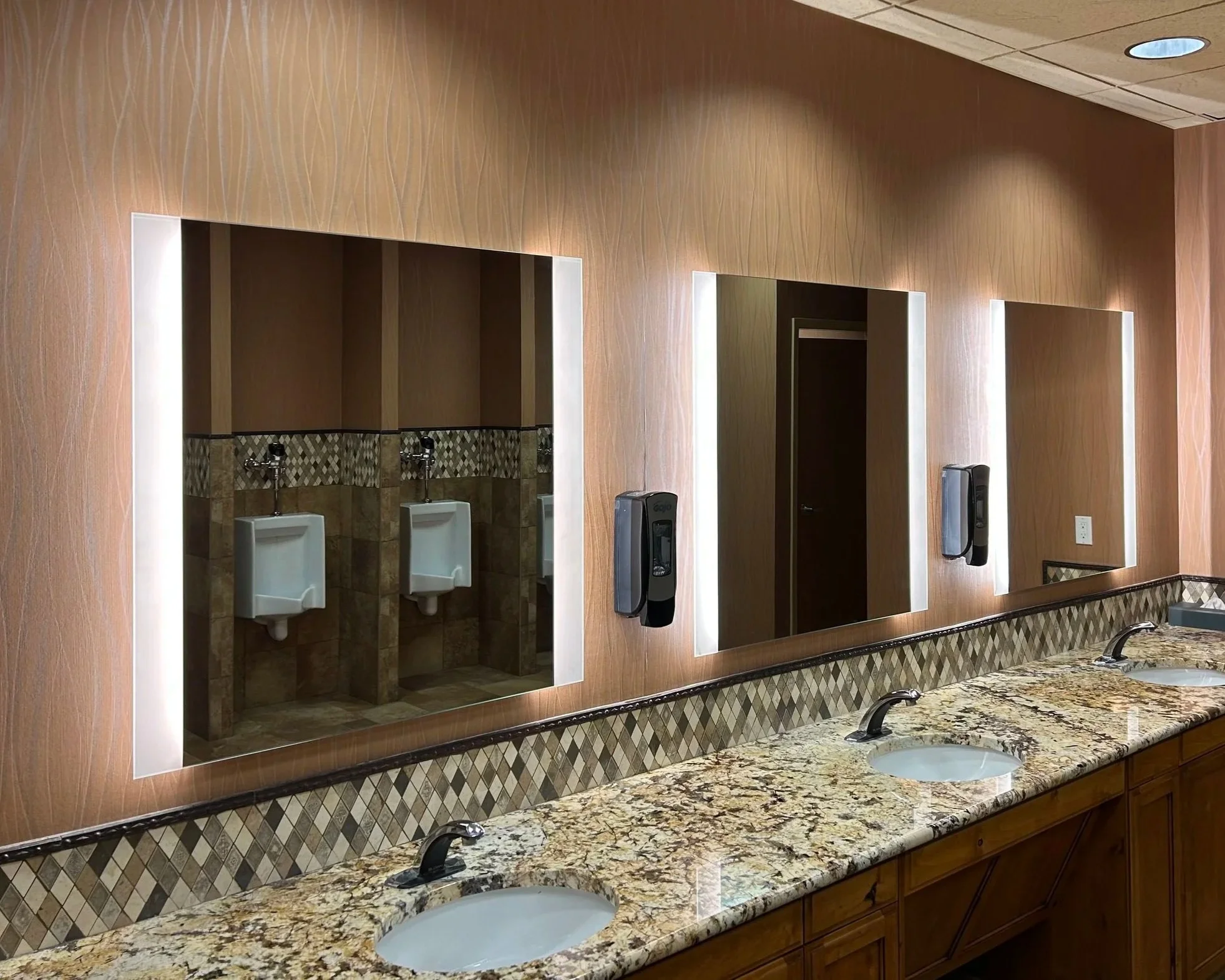Public restroom with three sinks, mirrors with backlighting, and urinals visible in the reflection of the left mirror.