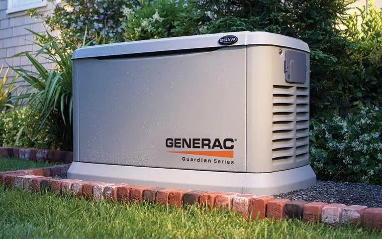 An outdoor Generac Guardian Series standby generator installed beside a garden bed with green plants and bushes.