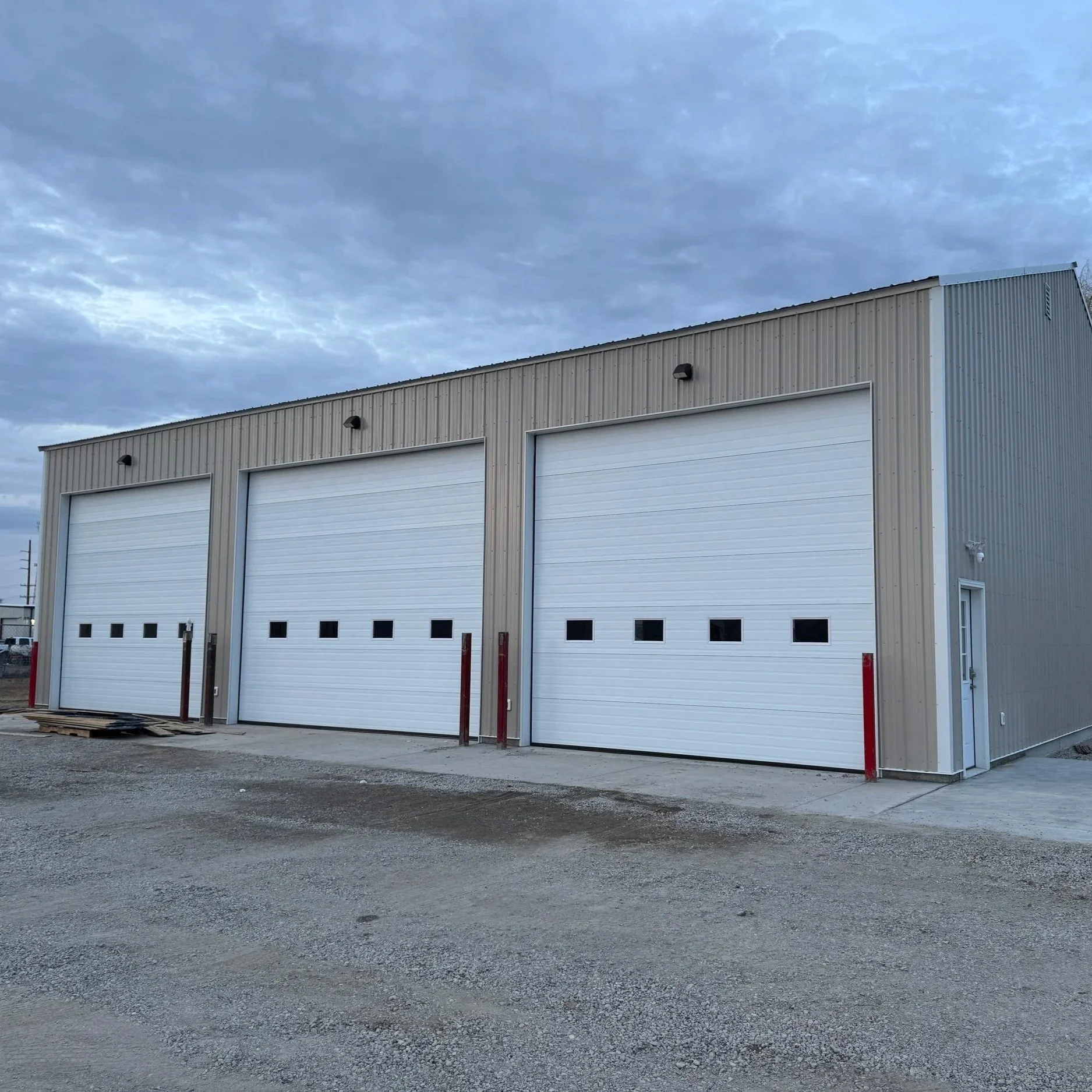 A large metal building with three white garage doors, situated in a gravel lot under a cloudy sky.