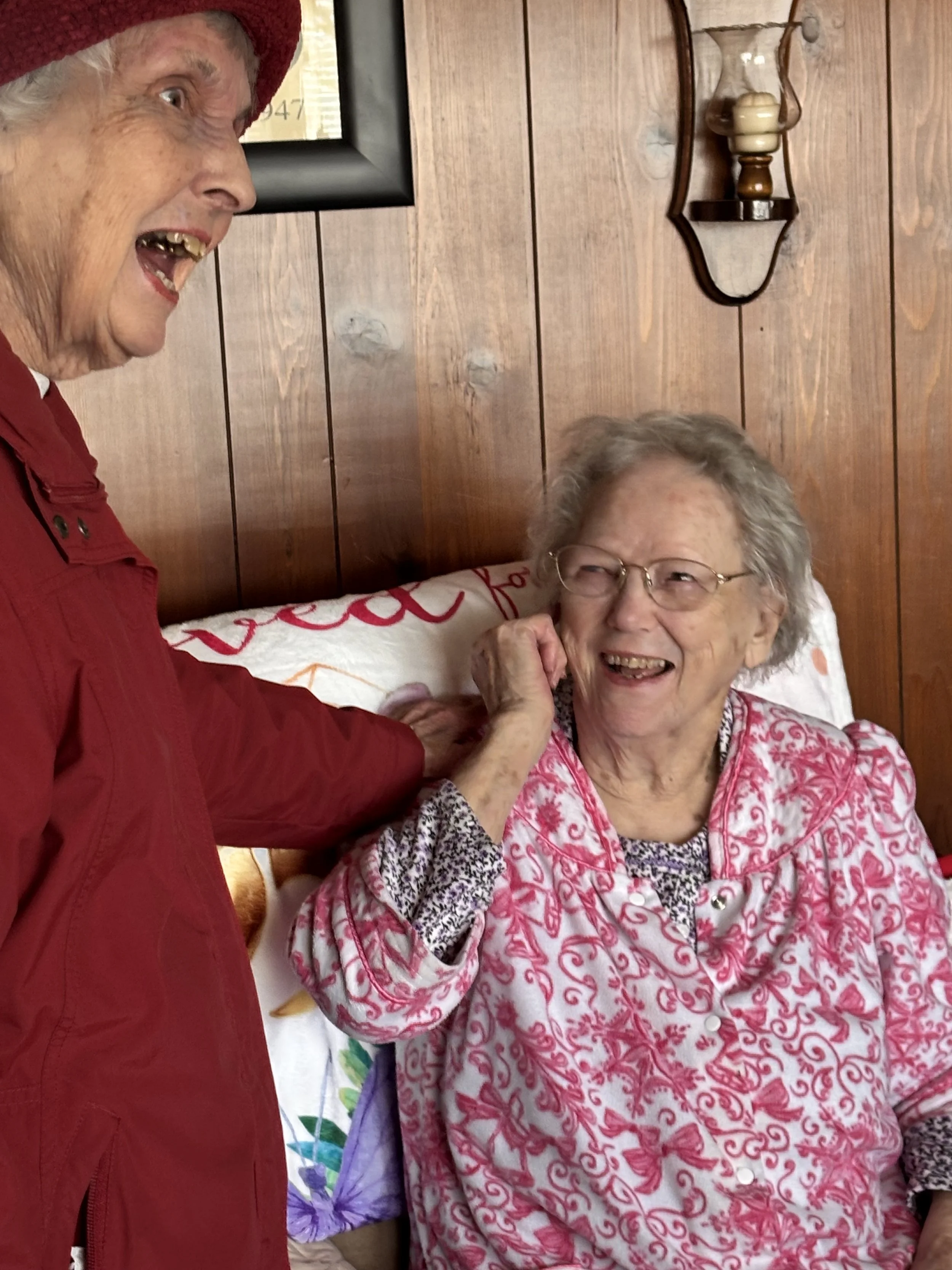 Two elderly women are sharing a happy moment, one lying in bed and the other standing beside her, gently touching her face. They are smiling and appear to be having a joyful conversation, with a wooden wall and decorative light fixture in the background.