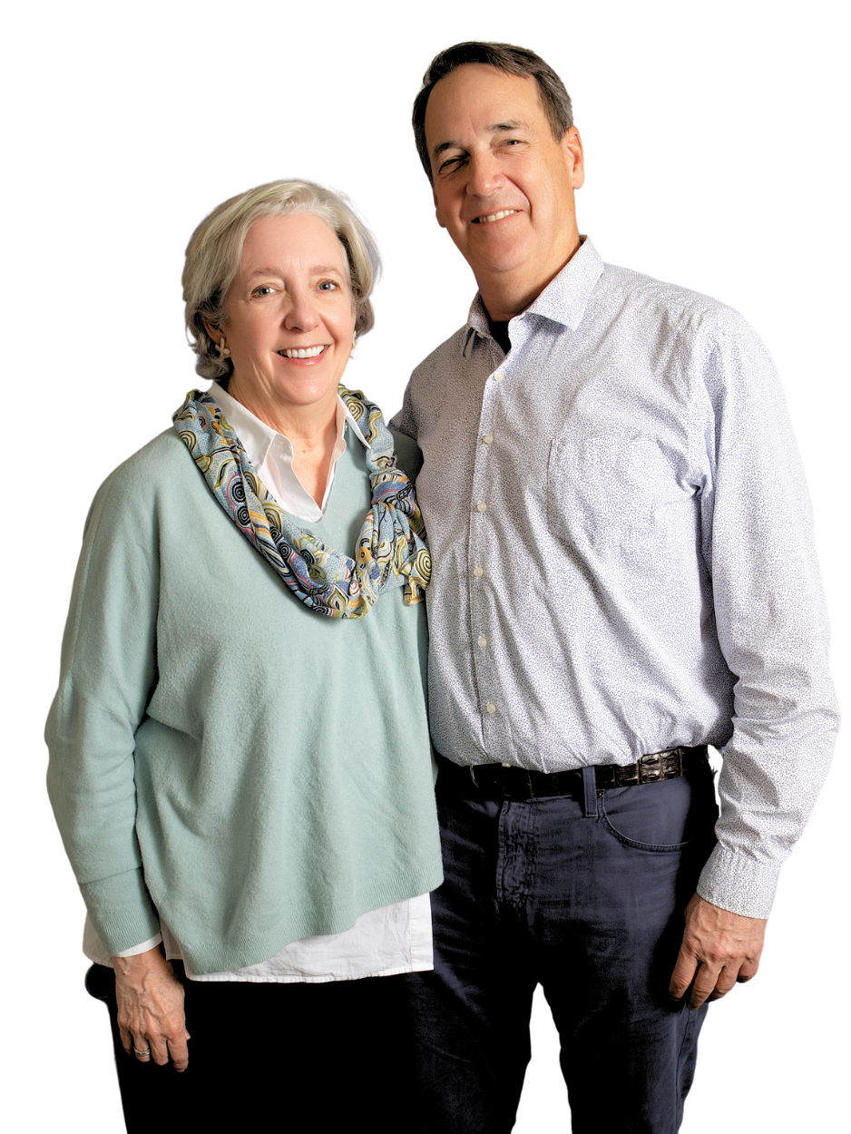 An elderly woman with short gray hair, smiling, wearing a light green sweater, white shirt, and a colorful scarf around her neck, standing next to a middle-aged man with short brown hair, smiling, wearing a light-colored patterned shirt, against a bl
