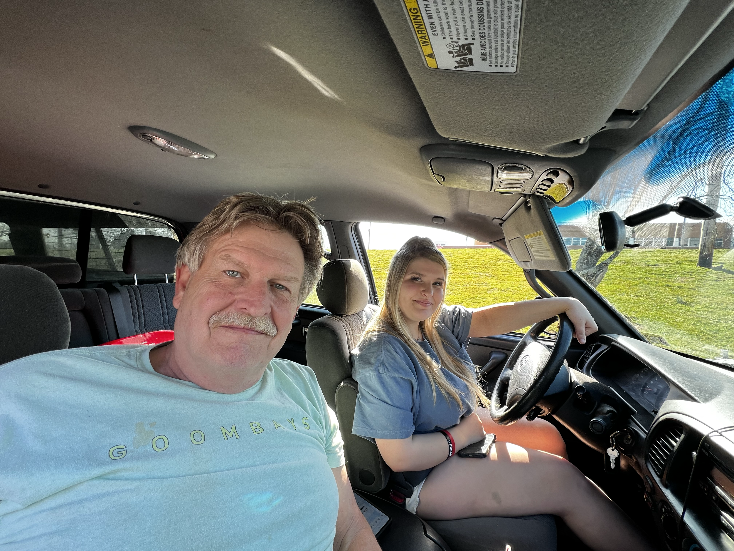 A middle-aged man with a mustache and a young woman with blonde hair sitting inside a vehicle, with the man in the foreground and the woman in the driver's seat, both smiling. Outside the vehicle, trees and a building are visible through the windows.