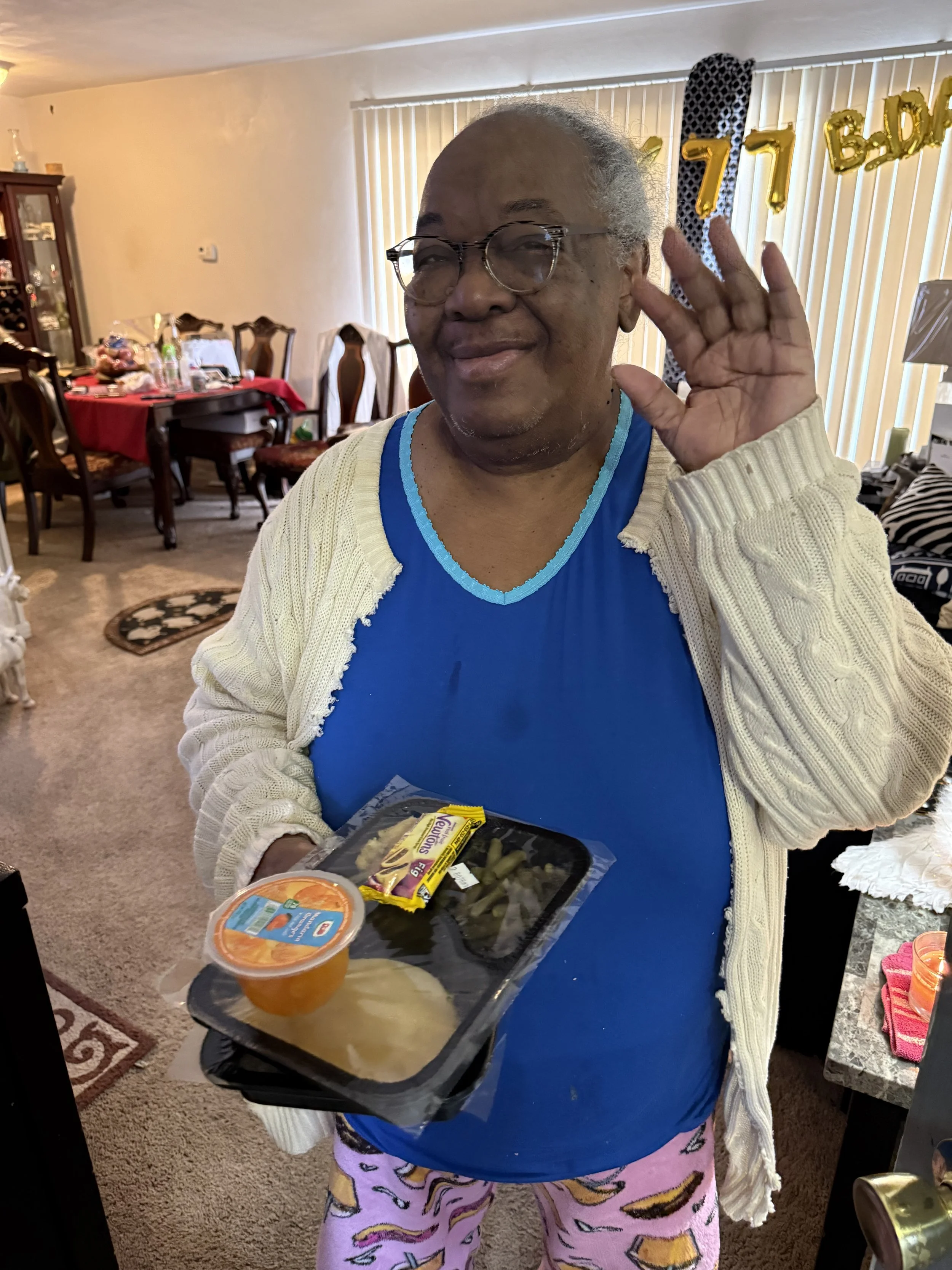 An elderly woman with glasses and a warm smile is waving her hand. She is wearing a blue shirt, a white cardigan, and pink pajama pants with a colorful pattern. She is holding a tray of food that includes a container of orange fruit, a bag of snacks,