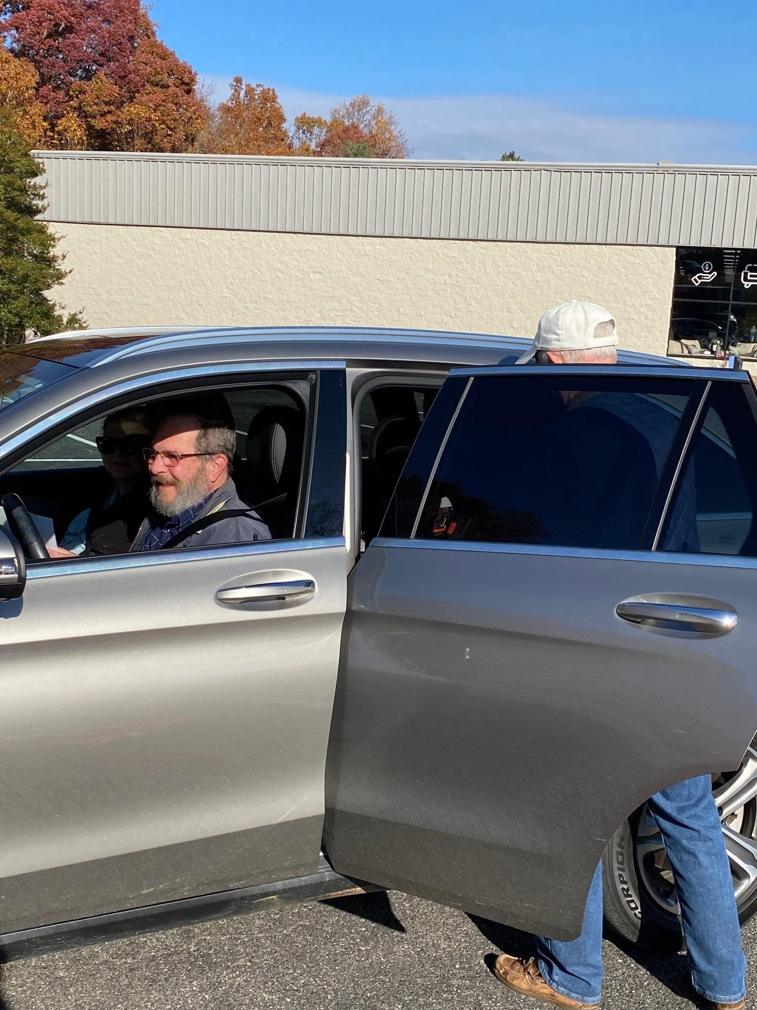 A man with glasses and a beard sitting in the driver's seat of a silver SUV, with a white cap on top of the vehicle. The vehicle is parked outside near a beige building with a textured wall and a sign with symbols, possibly indicating amenities. Trees with autumn-colored leaves are in the background, under a clear blue sky.