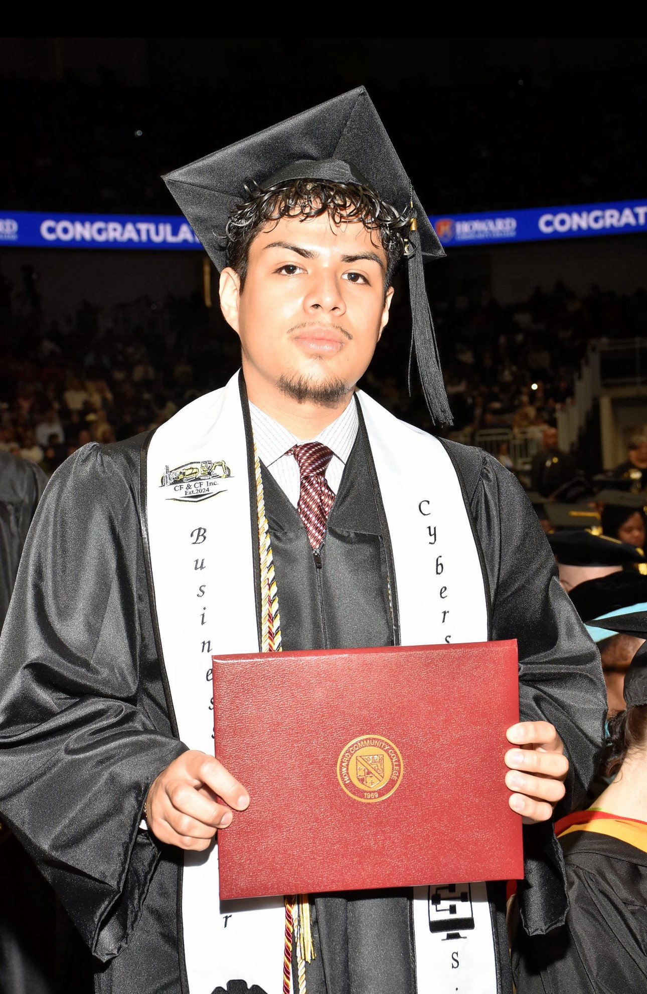A young man in a graduation cap and gown holding a diploma during a graduation ceremony at an arena.