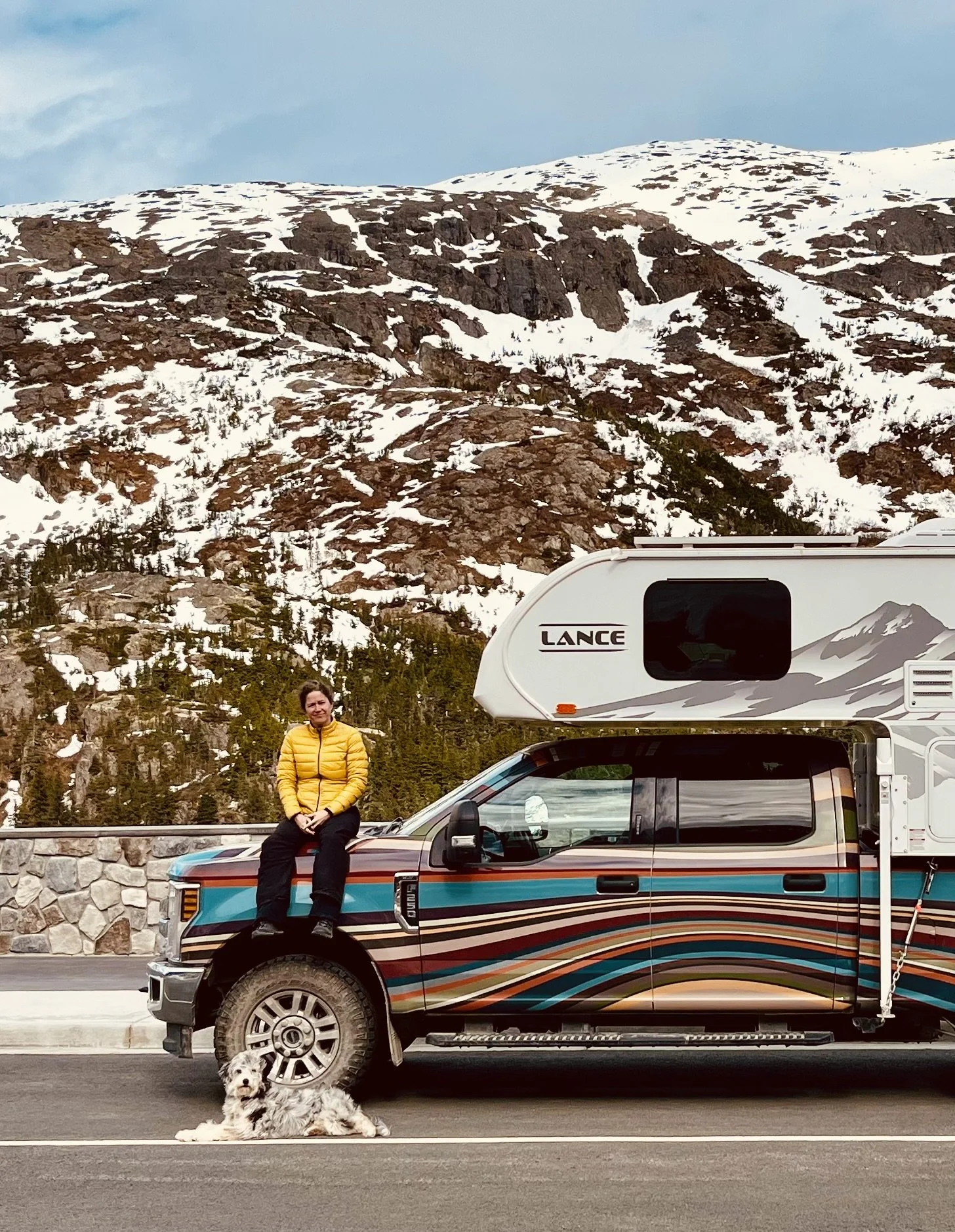 A woman sitting on the hood of a colorful pickup truck with a dog lying beside it, in front of a snow-covered mountain landscape.