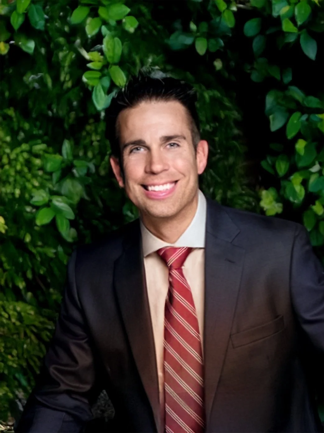 A man in a dark suit with a beige shirt and red striped tie smiling, standing in front of green foliage.