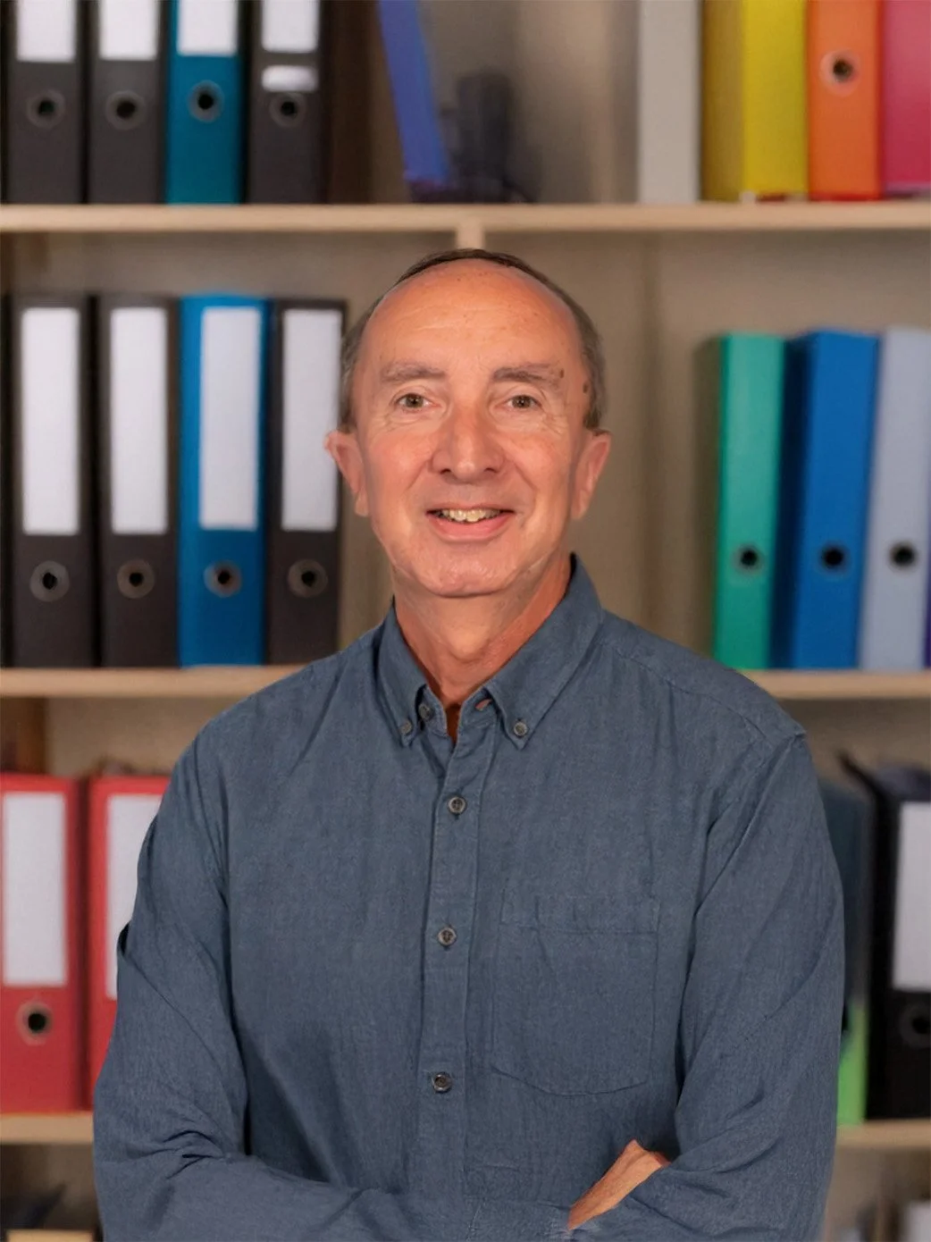 A man with a light blue button-up shirt smiling, standing in front of shelves filled with colorful binders.