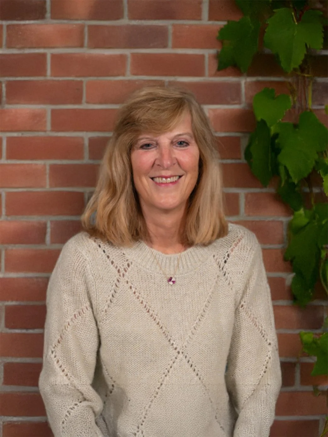 A smiling woman with shoulder-length blonde hair, wearing a beige knit sweater, standing in front of a red brick wall and green leafy vine.