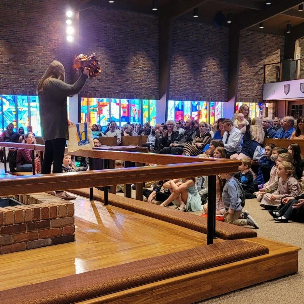 A woman standing on a wooden stage in a church, holding a bouquet of flowers and a gift bag, facing an audience of children and adults seated in pews. The church has stained glass windows and brick walls.