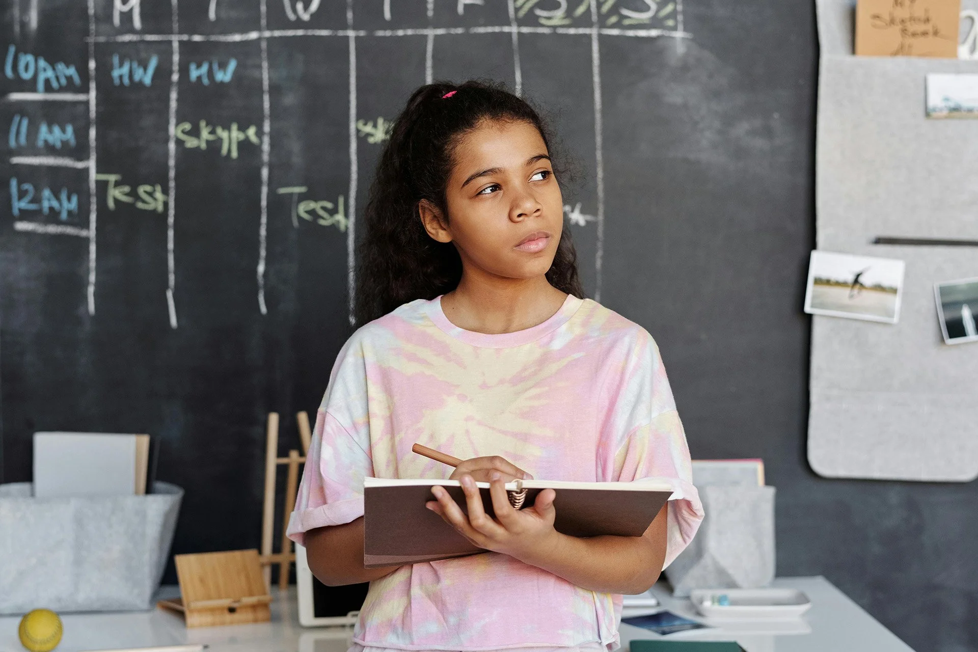 A young girl with curly dark hair and a pink hair clip, wearing a pastel pink and yellow tie-dye T-shirt, is standing in front of a black chalkboard with a schedule written on it. She is holding a spiral notebook and a pen, looking thoughtfully to the side.