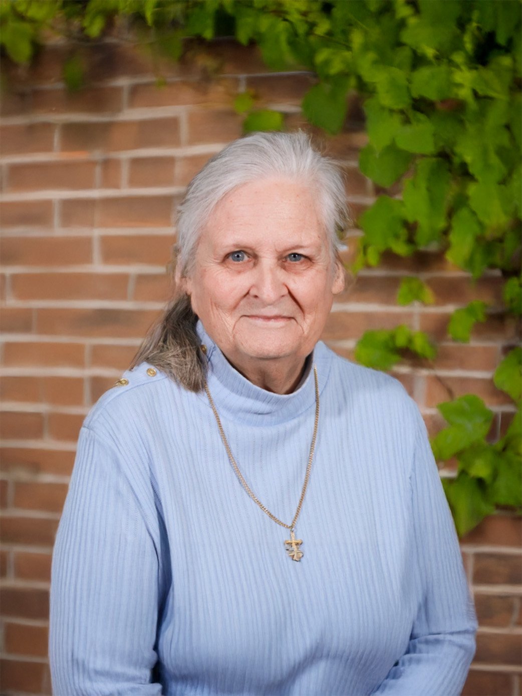 An elderly woman with gray hair, blue eyes, and light skin, wearing a light blue ribbed sweater and a gold necklace with a cross pendant, poses in front of a brick wall with green ivy.