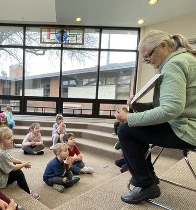 An older woman playing guitar while children sit on the floor, listening in a classroom with large windows and a stained glass window at the top.