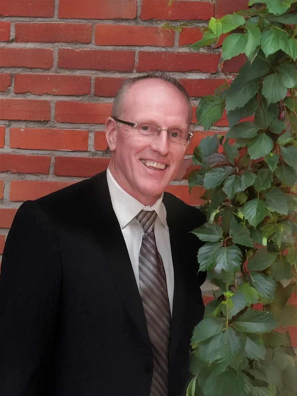 A man in formal attire with glasses, smiling, standing in front of a brick wall with green leafy vine growing on the right side.