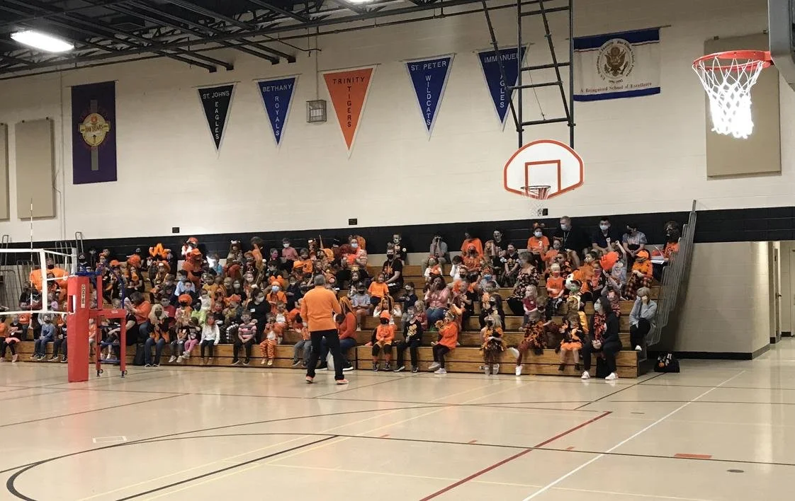 A gymnasium filled with children and adults wearing orange clothing, sitting on bleachers. The gym has banners hanging from the ceiling, a basketball hoop, and a volleyball net. Some people are wearing masks.