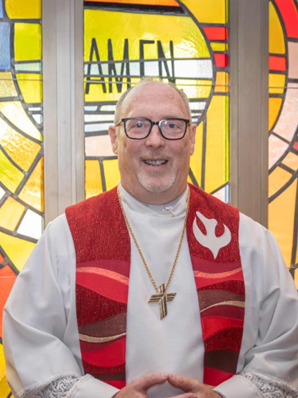 A smiling clergyman wearing glasses, a white robe, a red vestment with a flame symbol, and a gold chain with a cross pendant, standing in front of stained glass windows with yellow and orange colors.