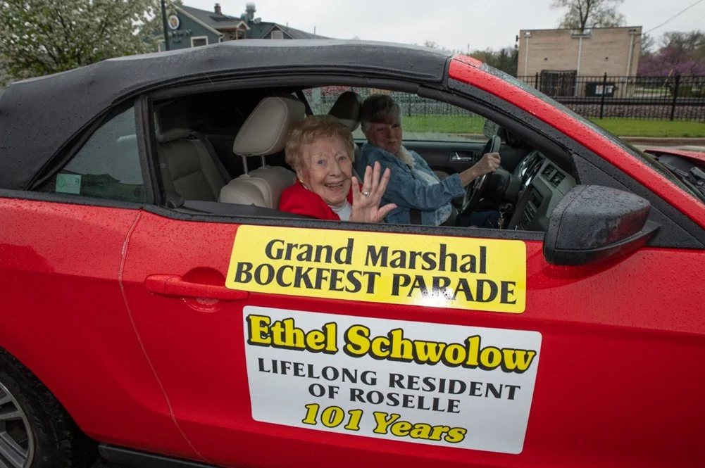 Two women inside a red convertible car with signs on the side. The signs read 'Grand Marshal Bockfest Parade' and 'Ethel Schwolow, Longtime Resident of Roselle, 101 Years.' The elderly woman in the passenger seat is smiling and waving, while the woman driving is looking forward. It is rainy outside.
