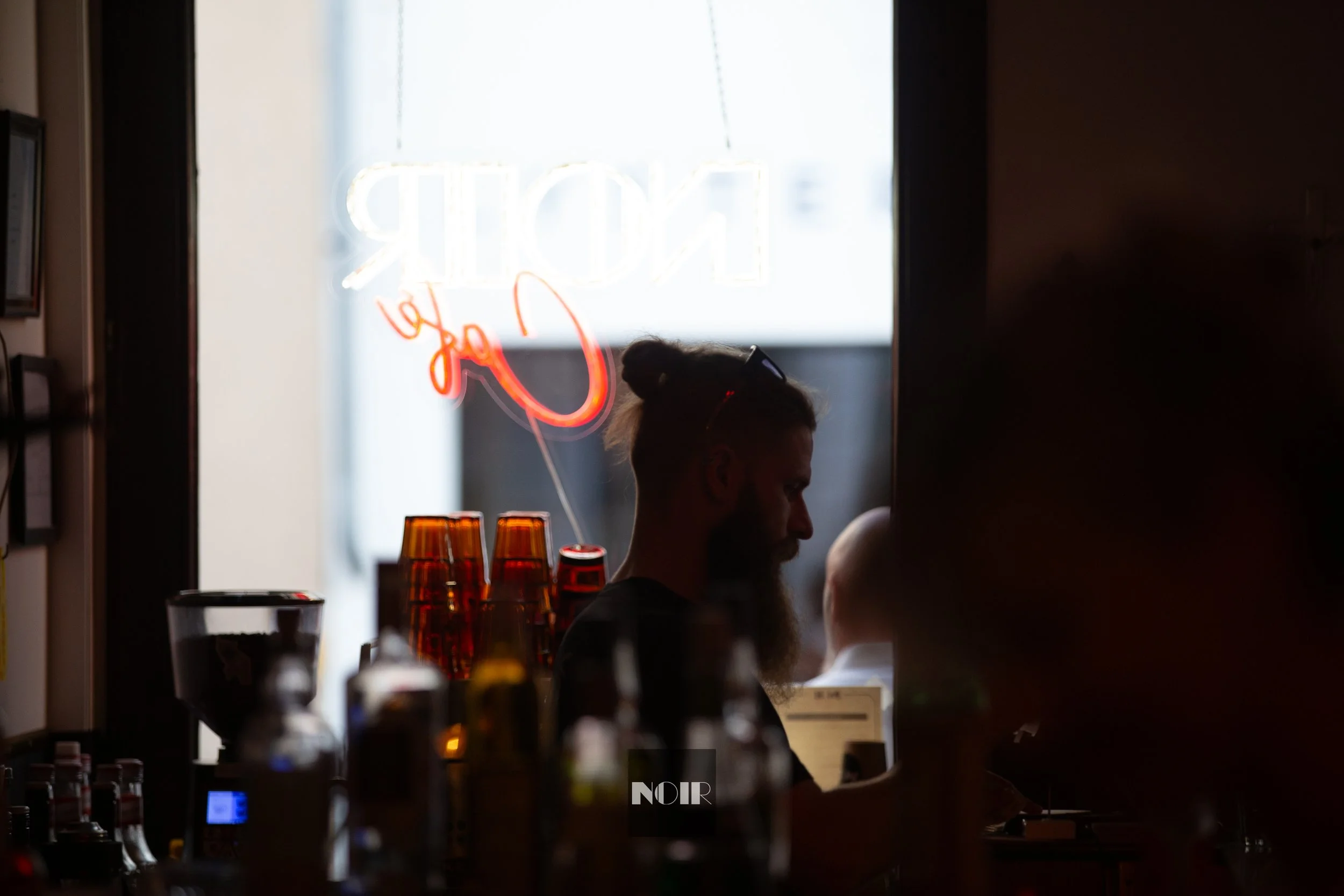 A person with a beard sitting at a bar or cafe, viewed through a window with a neon sign that reads 'ORDER by' in neon lights. The scene is dimly lit with bottles in the foreground.