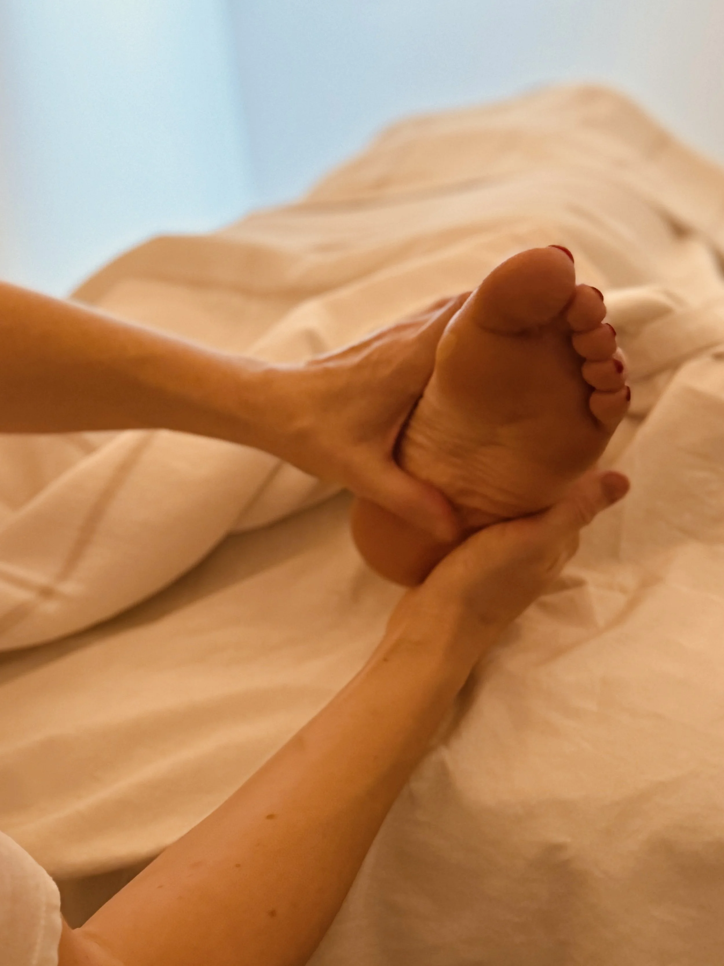 Close-up of a person holding an elderly person's foot in their hands, lying on a bed with light-colored sheets.