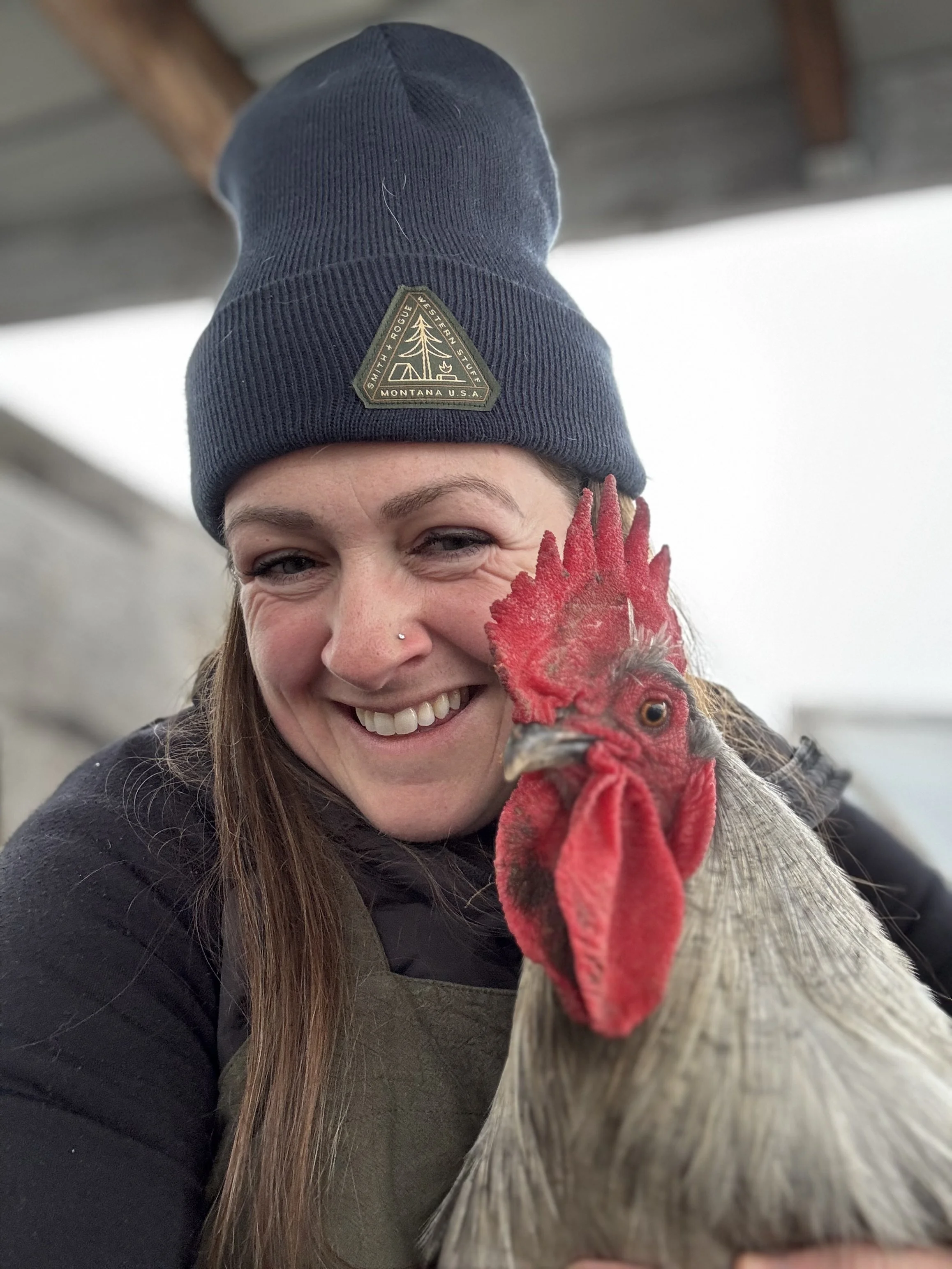 A woman smiling and holding a red rooster close to her face, wearing a gray beanie with a logo and a black jacket.