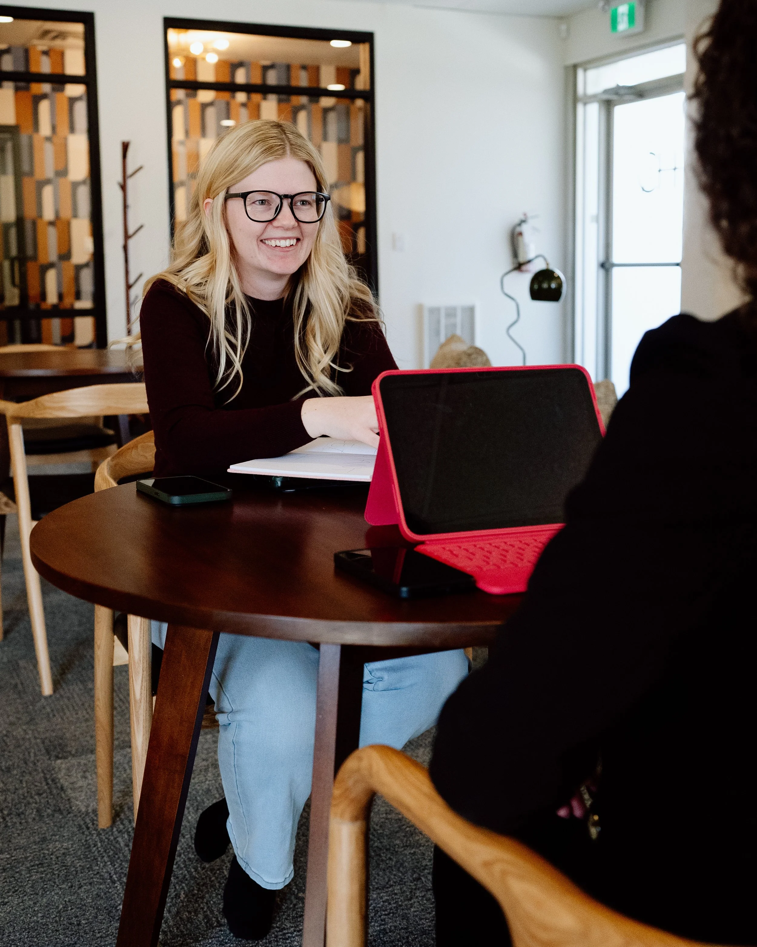 A woman with blonde hair, glasses, and a black top sitting at a round wooden table, smiling and talking to someone off-camera. There is a laptop with a red cover on the table.