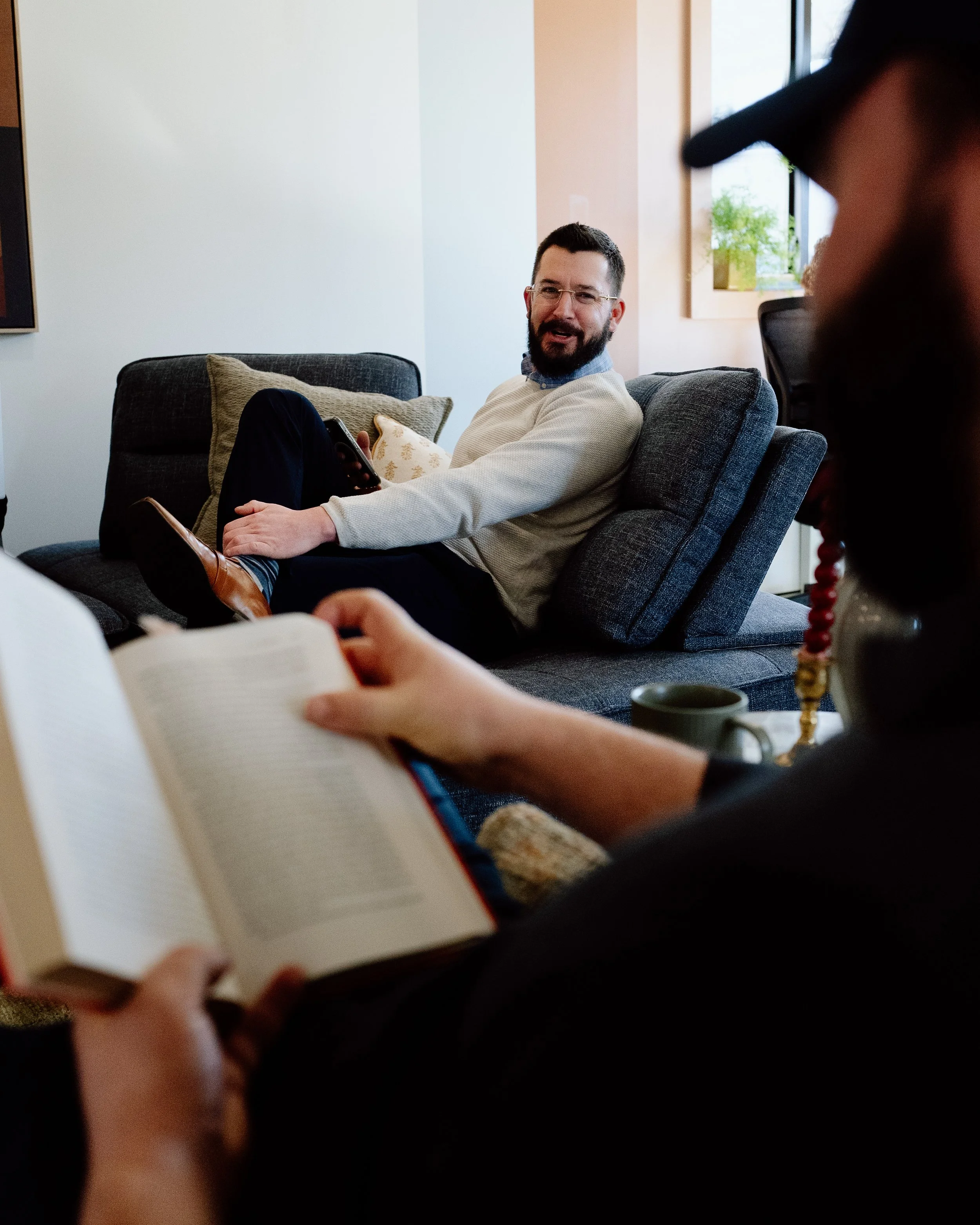 Man sitting on a gray sofa, holding a phone, smiling, talking to someone out of frame, in a cozy living room.