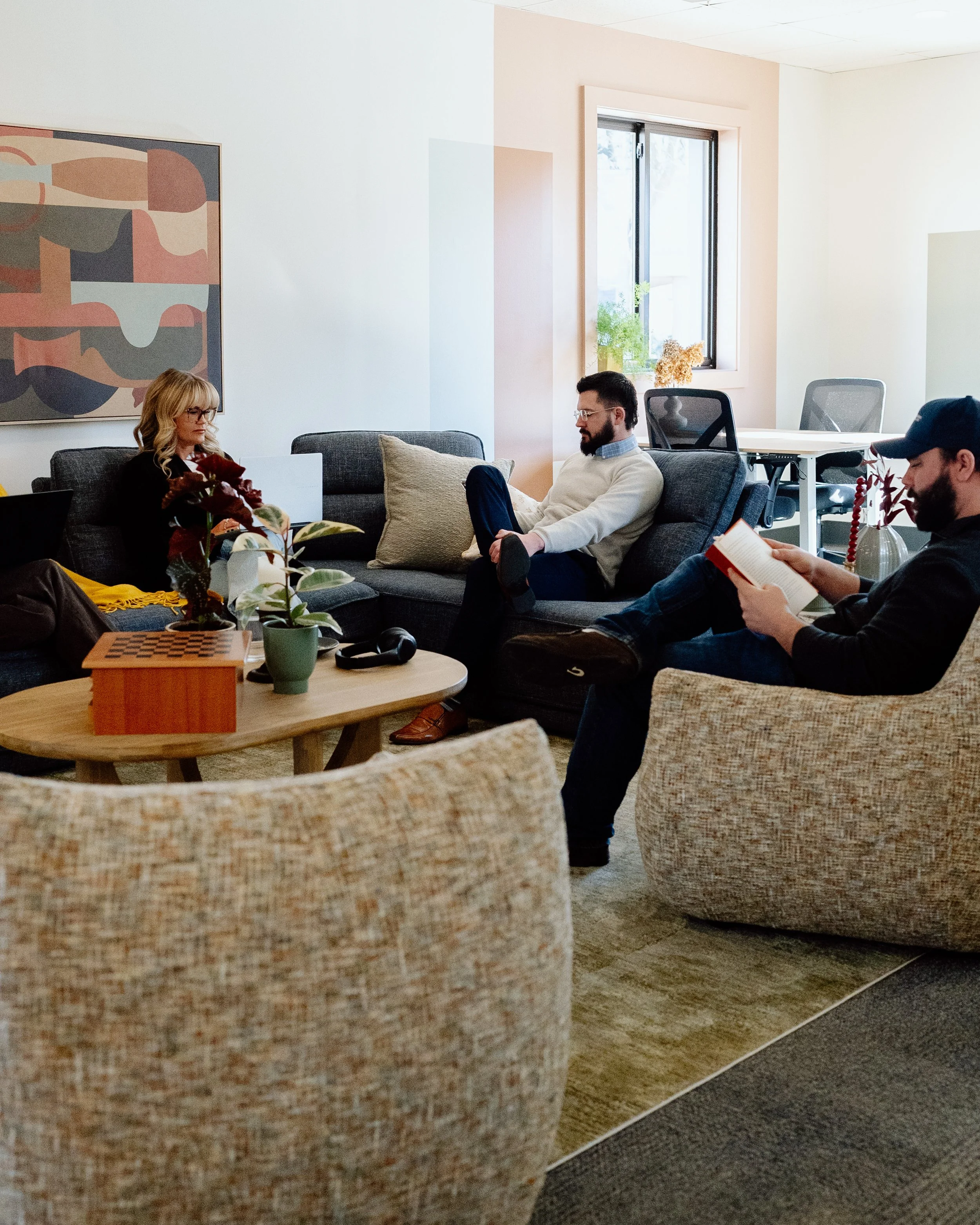 Three people sitting in a living room, two on a gray sofa and one in a beige armchair, engaged in different activities. The woman on the sofa is sitting with a laptop, the man next to her is sitting on the same sofa looking at his phone, and the man in the armchair is reading a book. There are plants and artwork on the walls, with sunlight coming in through a window.