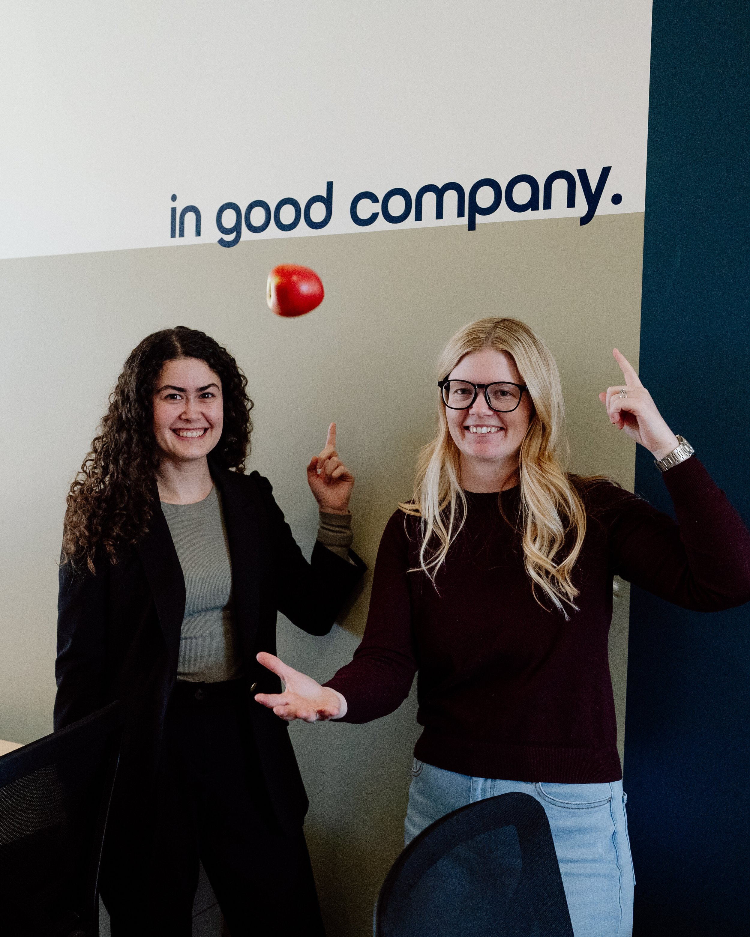 Two women smiling and pointing upwards, with an apple in mid-air between them, in front of a wall with the words 'in good company'.