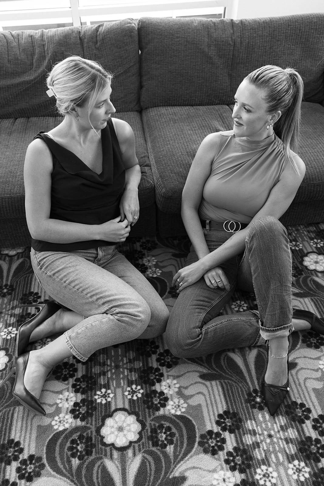 Two women kneeling on a floral rug, facing each other, engaged in conversation, with a couch behind them.