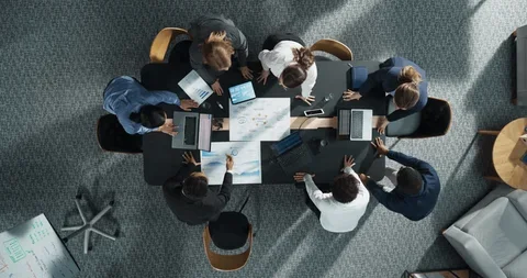 A group of eight people sitting around a rectangular conference table with laptops and tablets, discussing and working collaboratively.