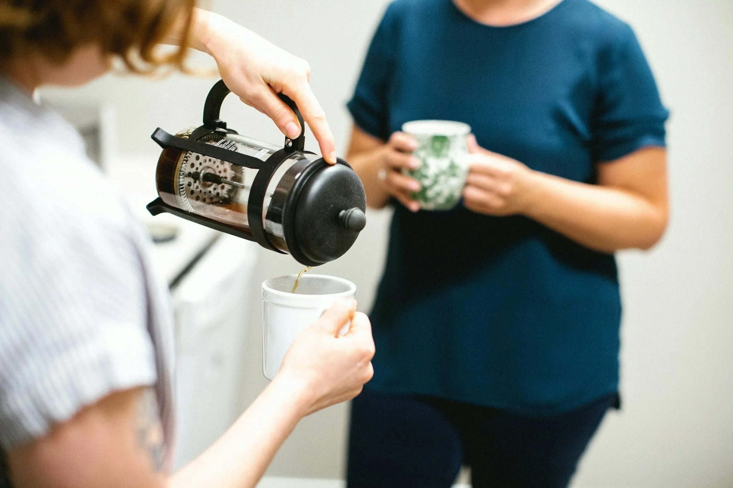 A person pours coffee from a French press into a white mug while another person holds a green patterned mug in the background.