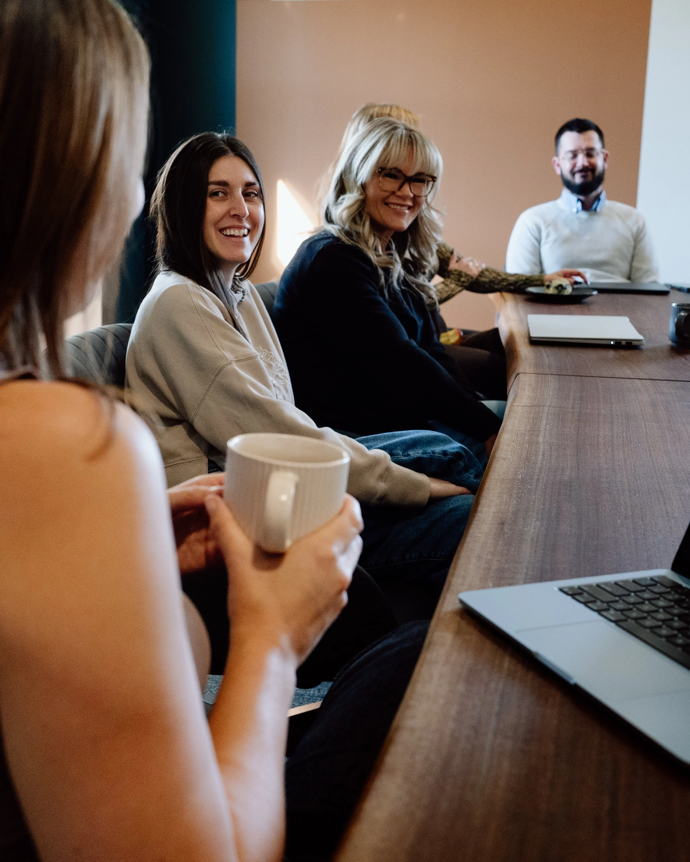 Group of people sitting at a conference table, smiling and engaging in a meeting or discussion, with laptops and coffee mugs on the table.