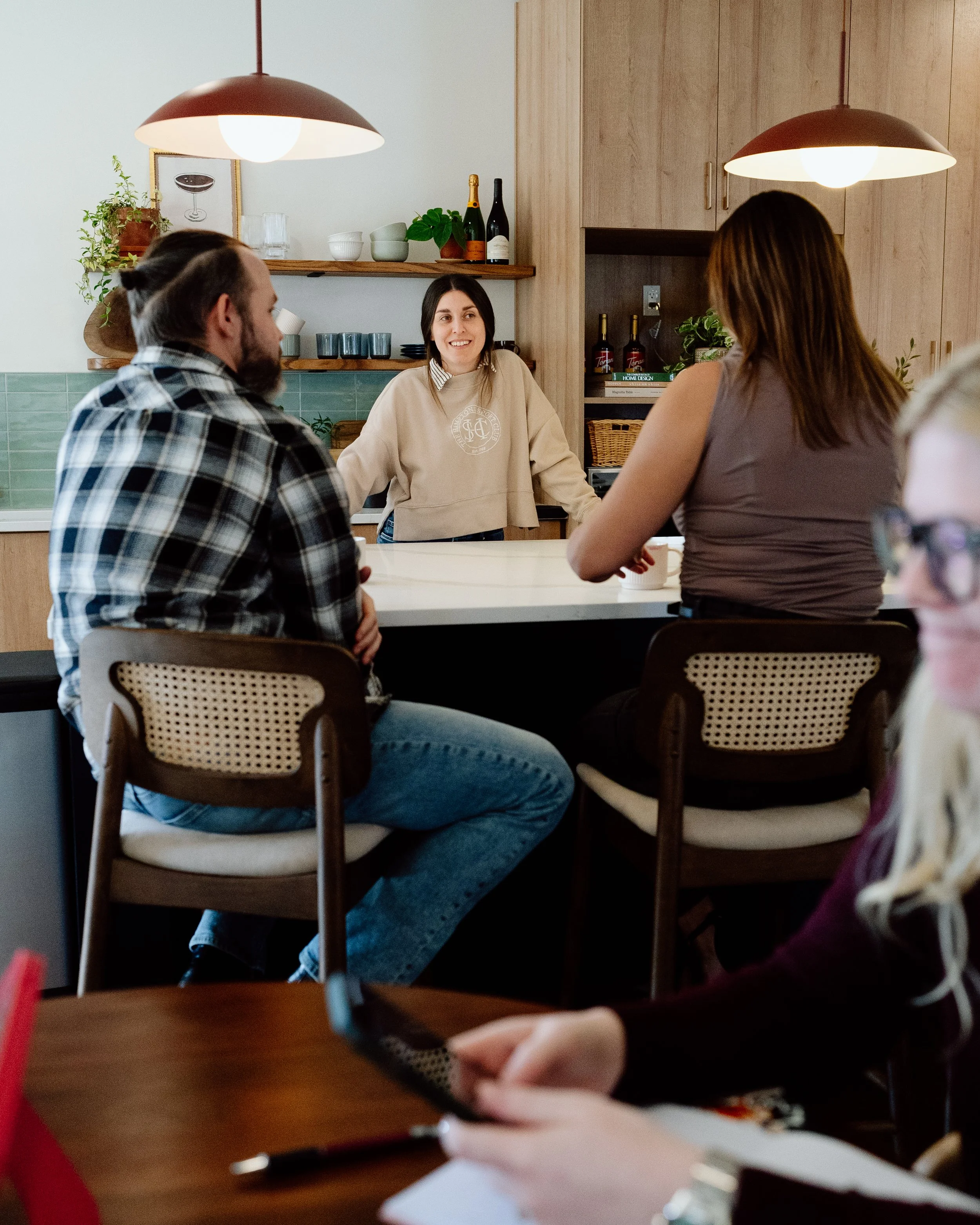 People sitting at a kitchen bar having a conversation, with a woman standing behind the counter smiling.