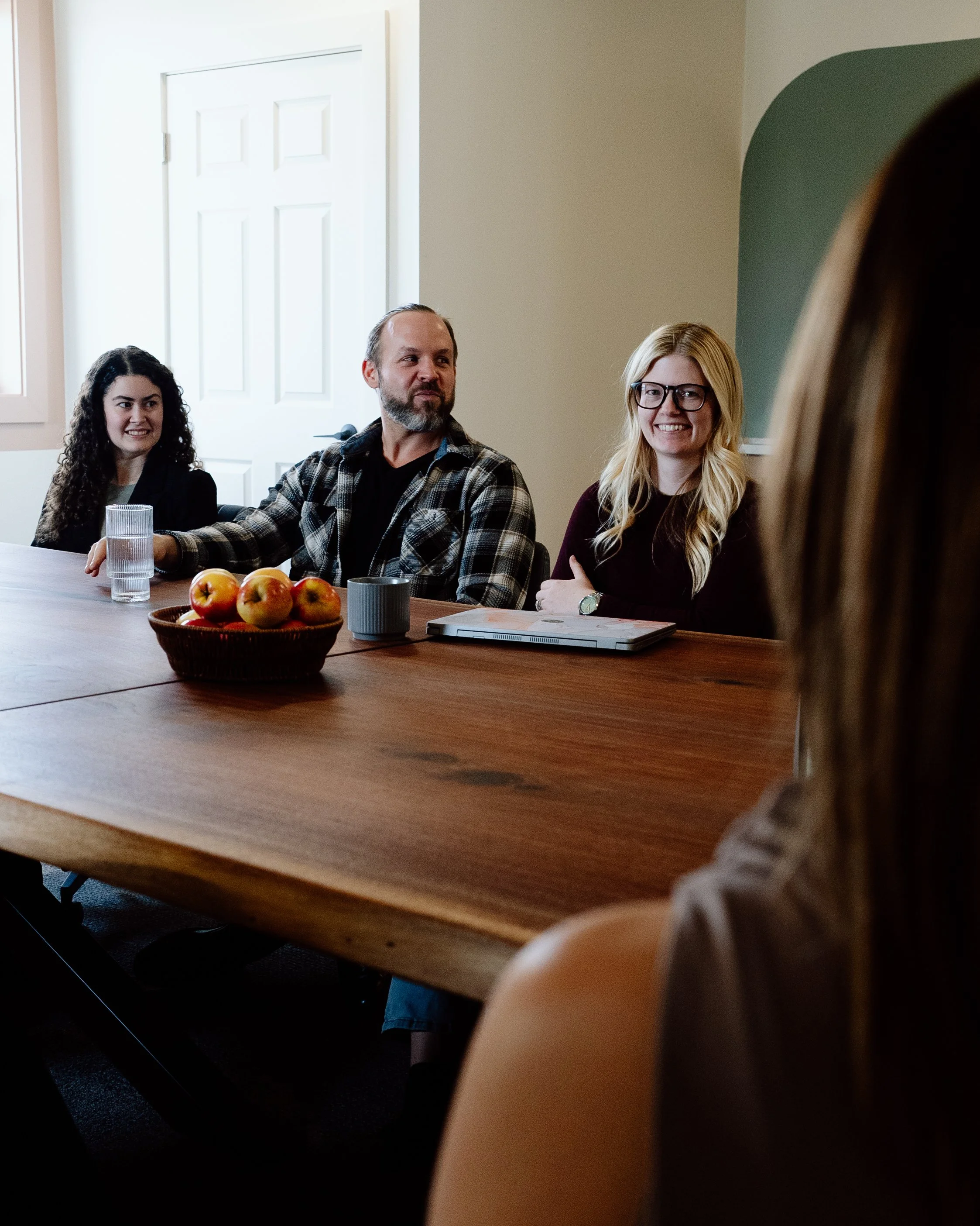 People sitting around a conference table in a meeting room, with a bowl of apples and drinks. One woman faces away from the camera, while a man and two women look towards her, smiling.