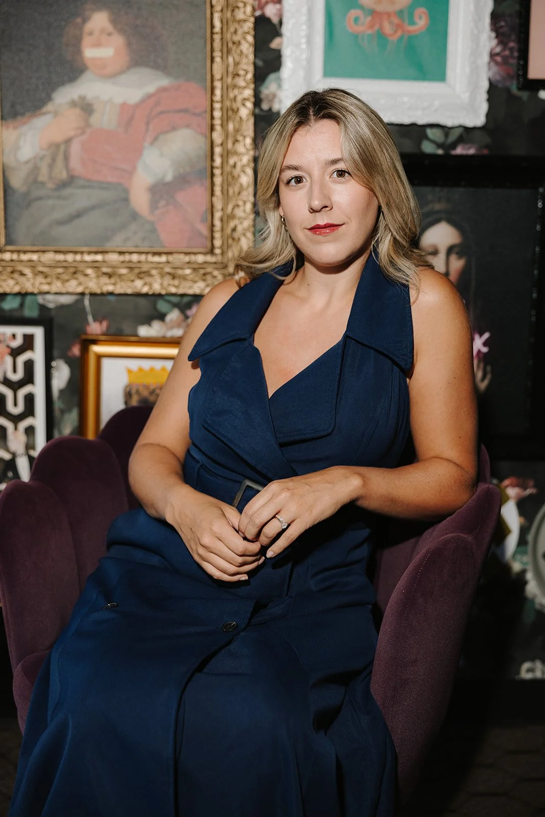 A woman with shoulder-length blonde hair wearing a sleeveless dark blue dress, seated on a purple chair in front of a dark, decorated wall with framed artwork and photographs.