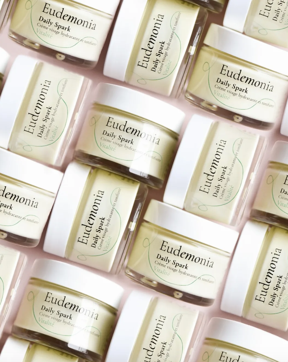 Multiple jars of Eudemonia Daily Spark moisturizing cream arranged in a pattern on a pink surface.