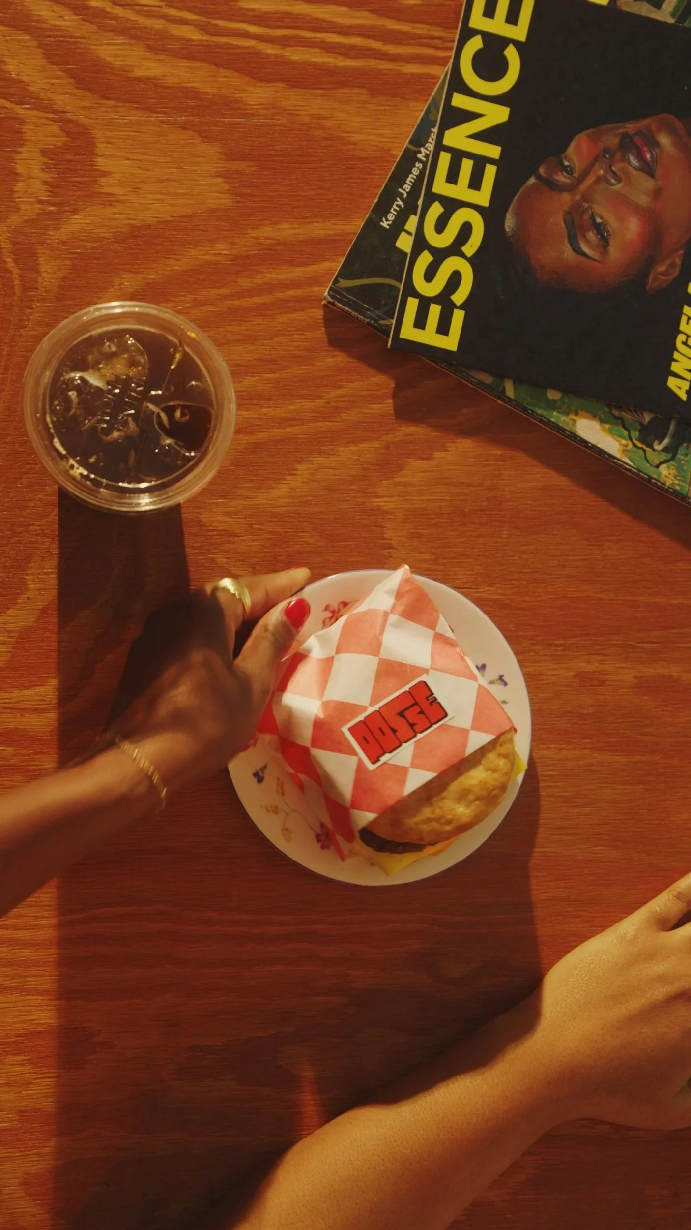 A hand with red nail polish and gold jewelry reaching for a wrapped Egg McMuffin breakfast sandwich on a white plate with a red and white checkered wrapper, on a wooden table. A plastic cup of soda with ice and a book titled "Essen" featuring a smiling woman on the cover are also on the table.