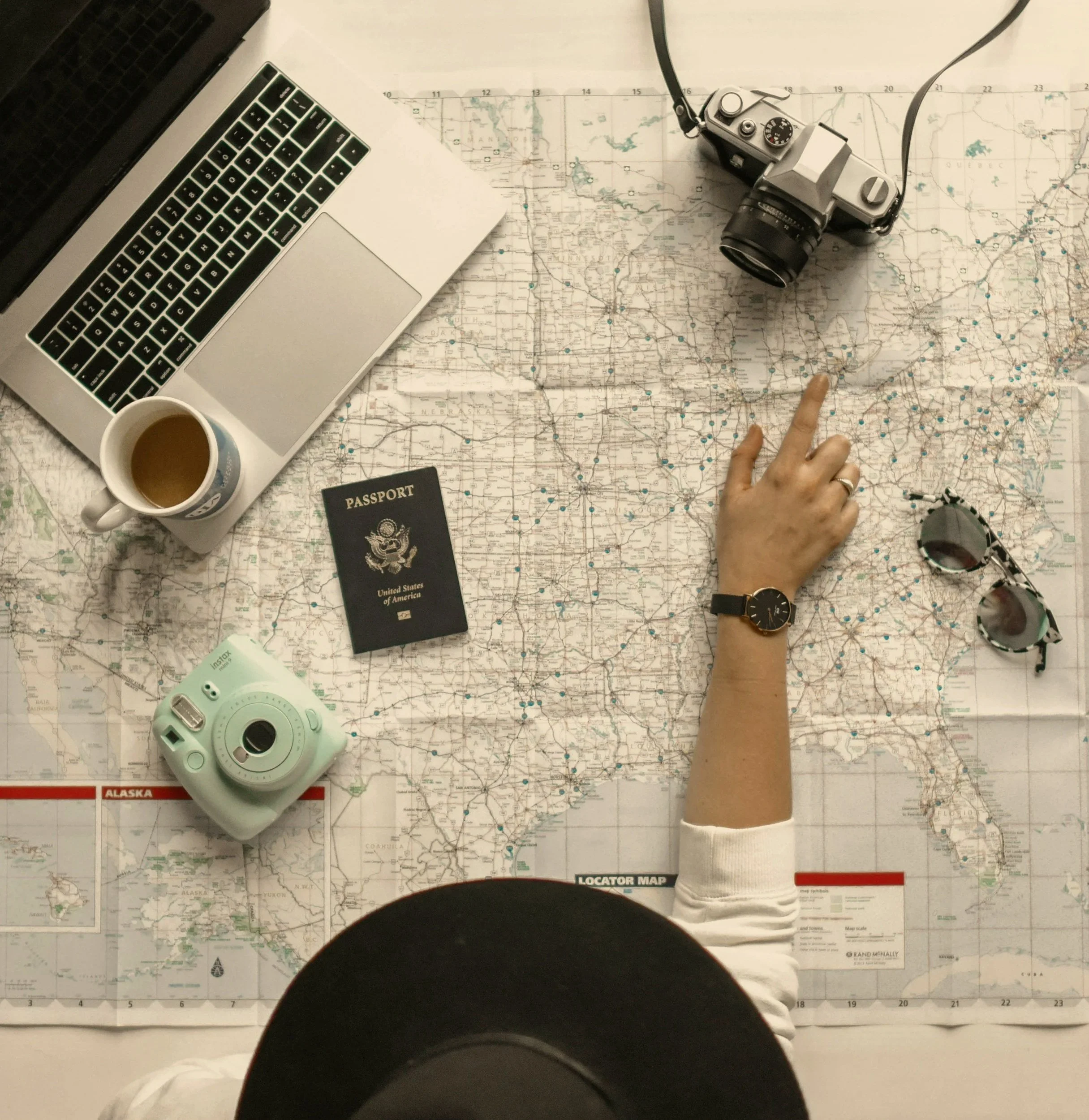 Top-down view of a table with a laptop, a map, a camera, a cup of coffee, a U.S. passport, and a locator map.