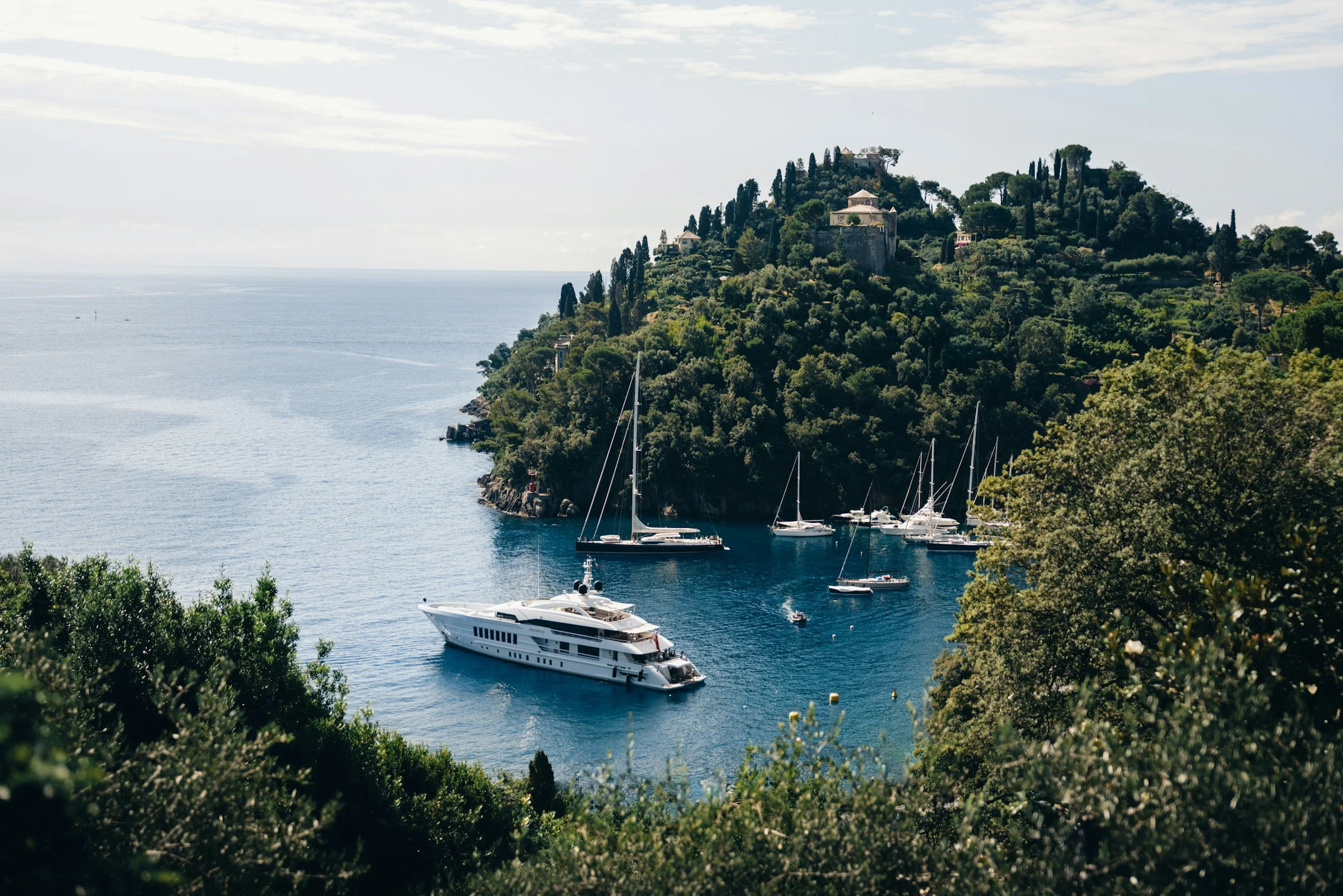 A coastal scene with a hillside covered in lush green trees and buildings, overlooking a harbor with multiple sailboats and a large luxury yacht, under a partly cloudy sky.