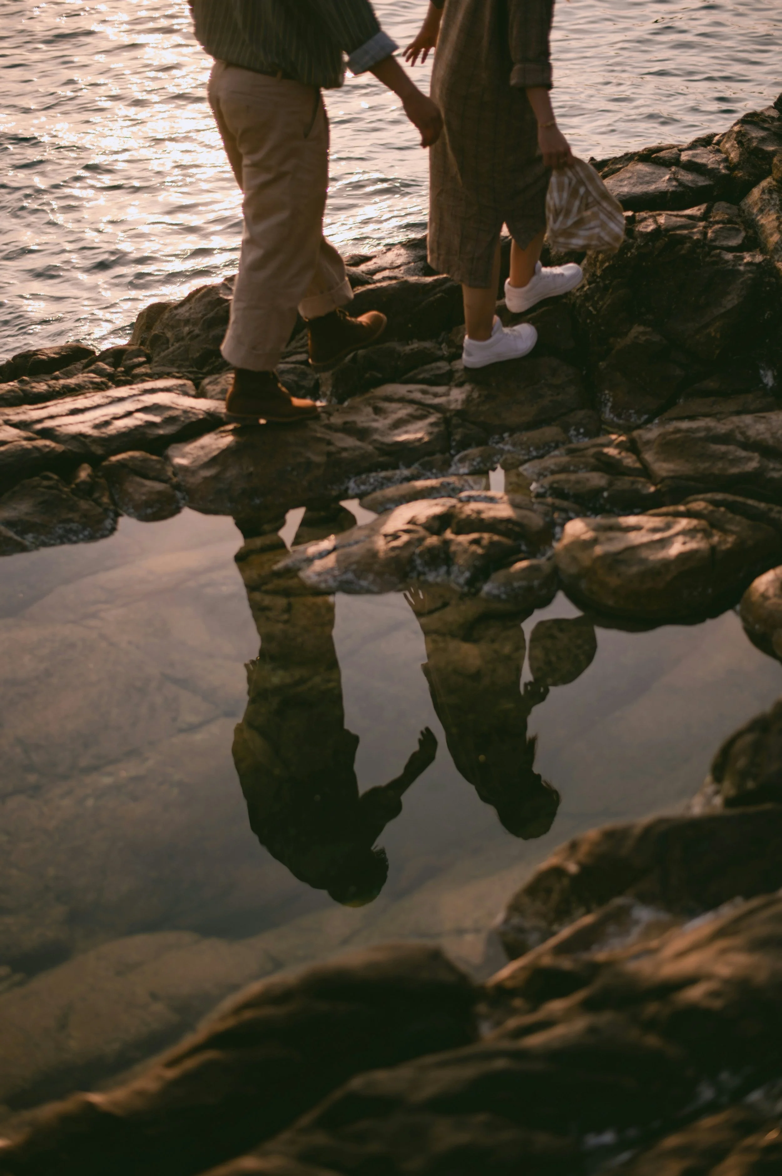 A couple standing on rocks by the water during sunset, holding hands and with their reflection visible in the water.