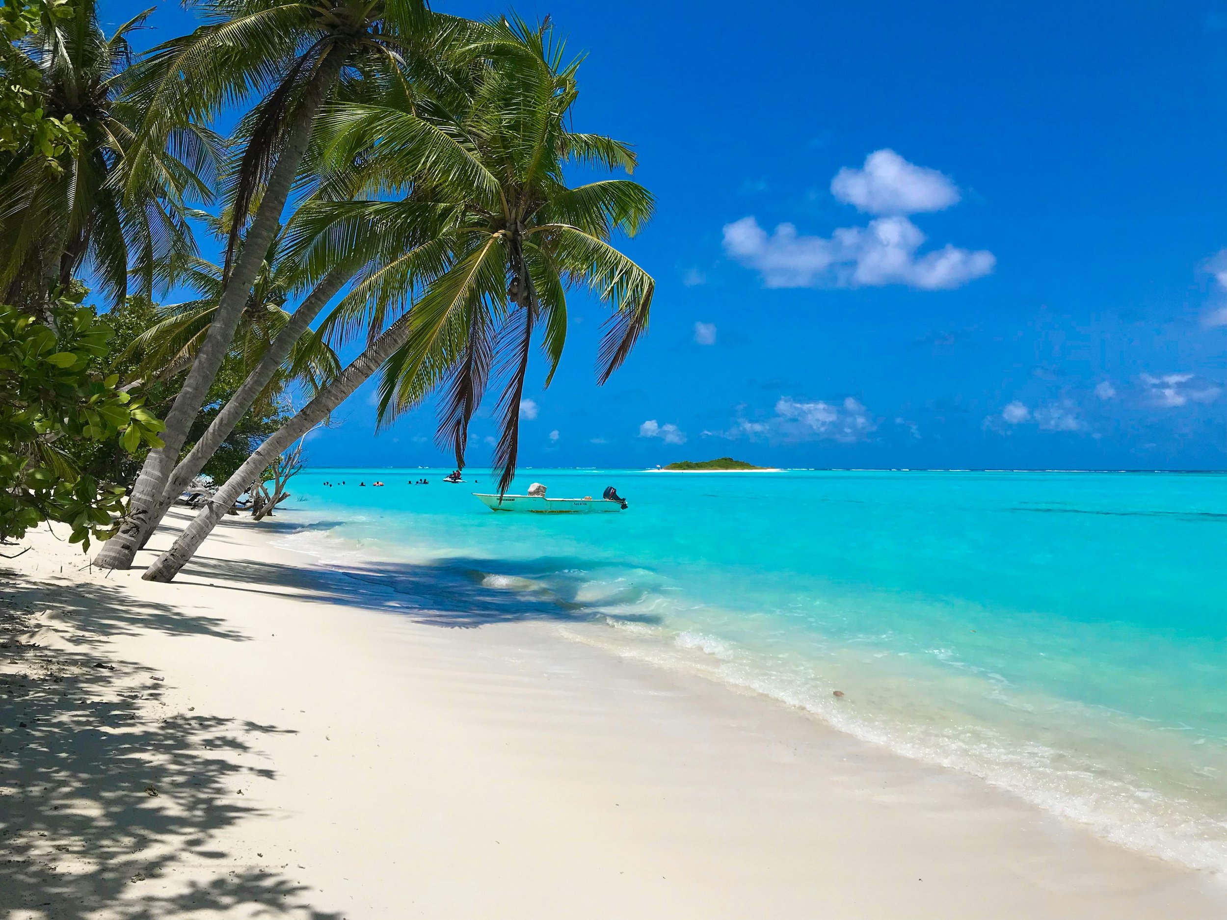 Tropical beach with white sand, turquoise water, palm trees, and a small island in the distance under a blue sky with some clouds.