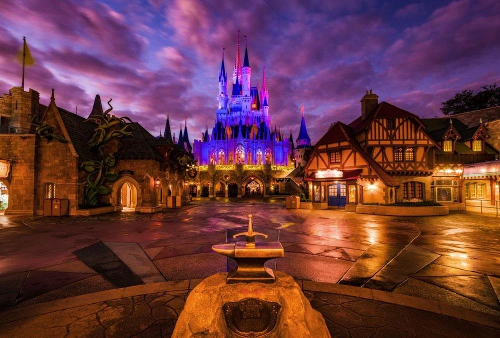 Disney World's Cinderella Castle illuminated with purple and pink lights at dusk, surrounded by themed buildings with fantasy architecture.