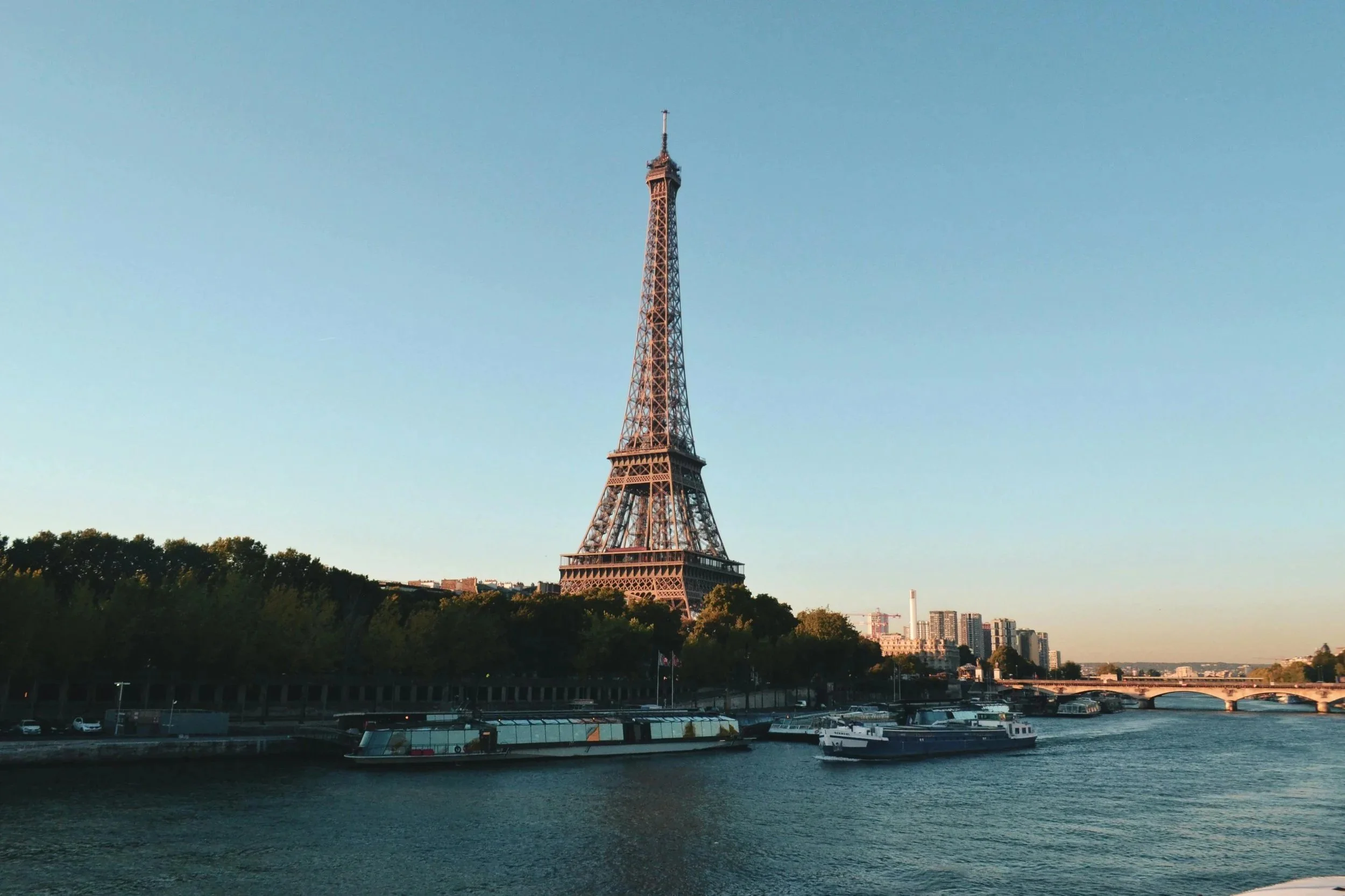 The Eiffel Tower standing tall over the river and cityscape in Paris, France, with boats on the river and greenery in the foreground.