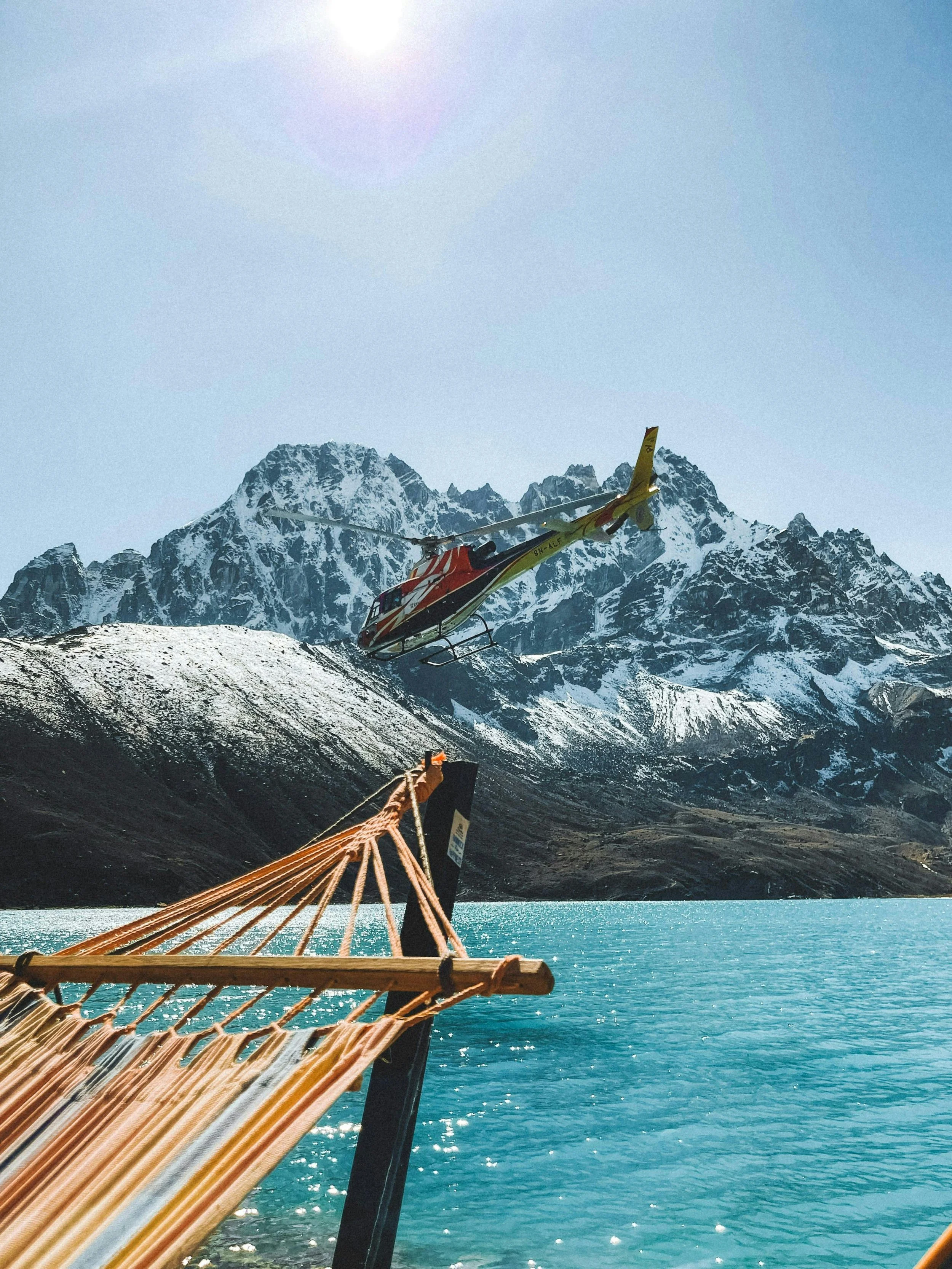 A helicopter flying over snow-capped mountains near a lake, with part of a rope hammock in the foreground.