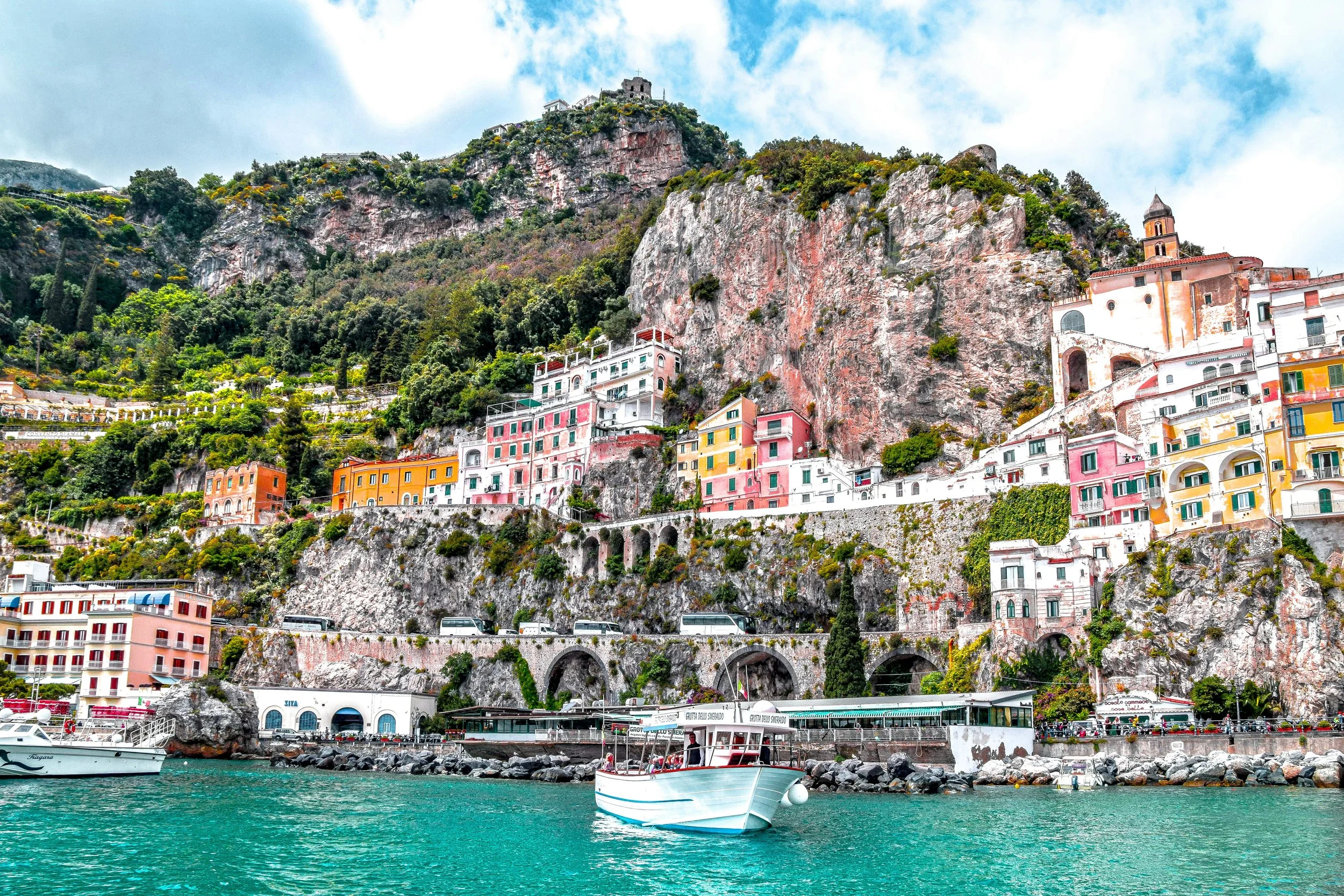 Colorful buildings on a hillside overlooking a harbor with boats, set against rocky cliffs and a partly cloudy sky.