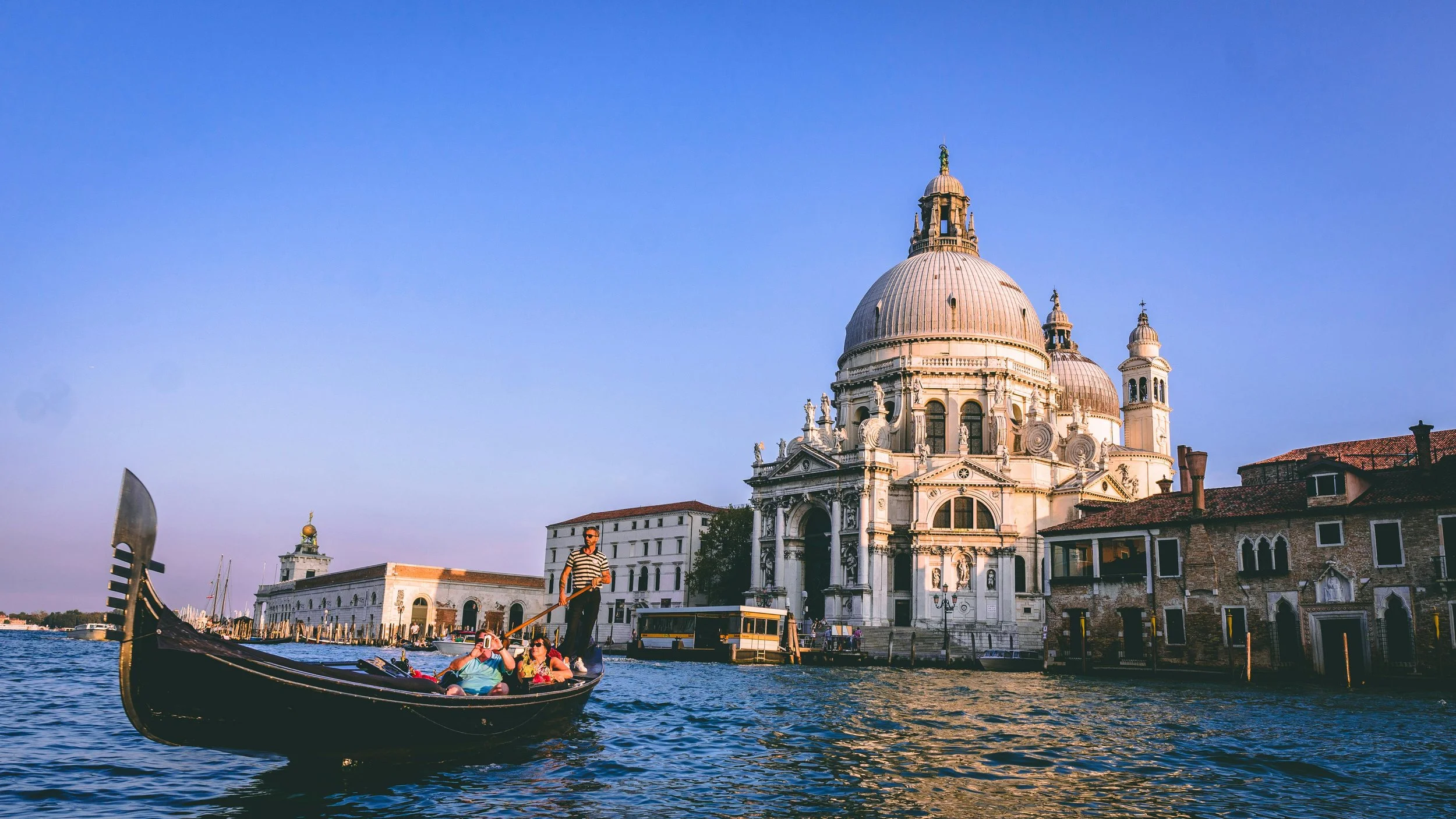 A gondola with tourists on the water in Venice, Italy, with historic buildings and a large domed church in the background under a clear blue sky.