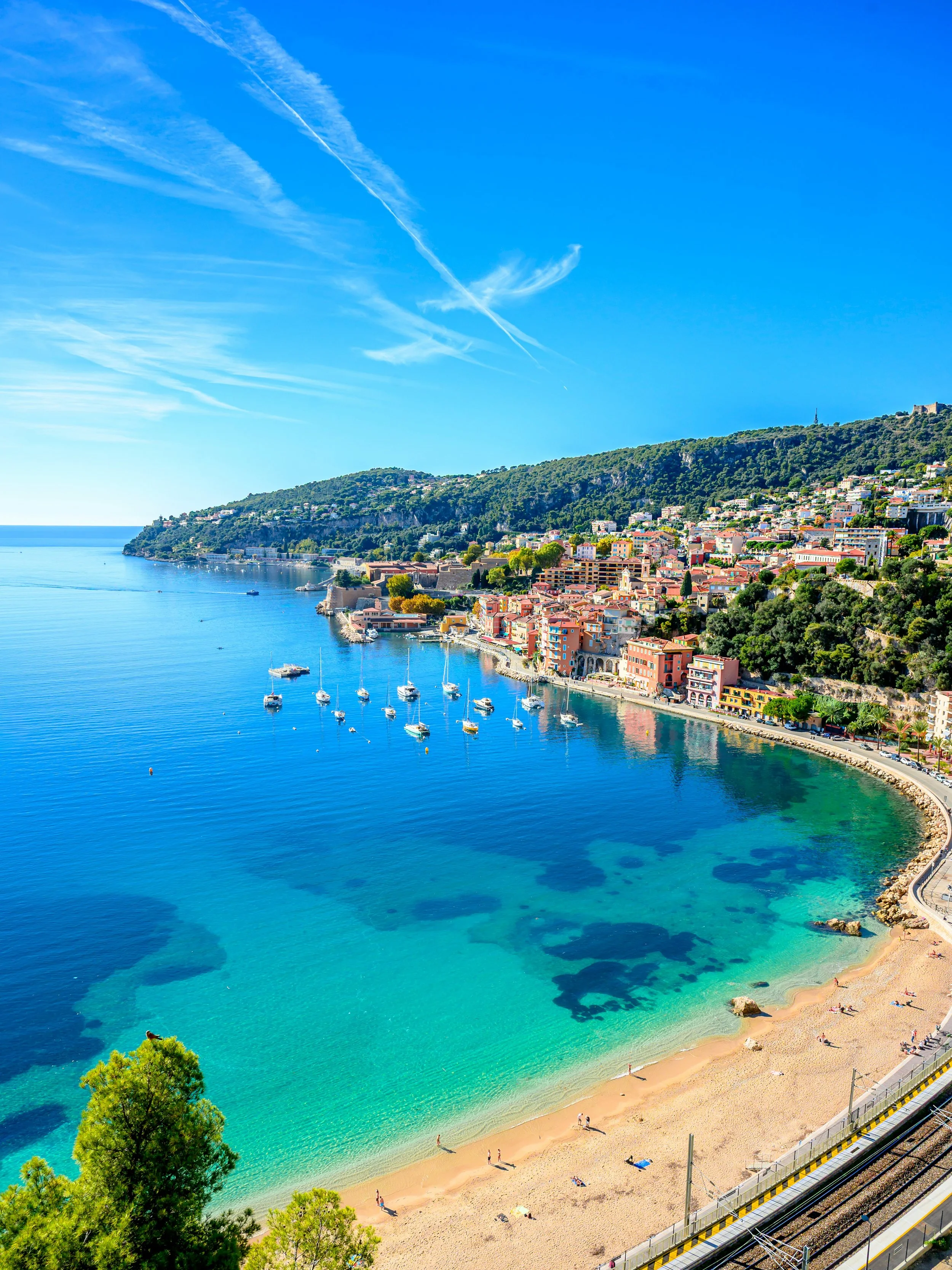Coastal town with colorful buildings on a hillside, clear turquoise water, sandy beach with people, trees in the foreground, and a railway track running along the coast.