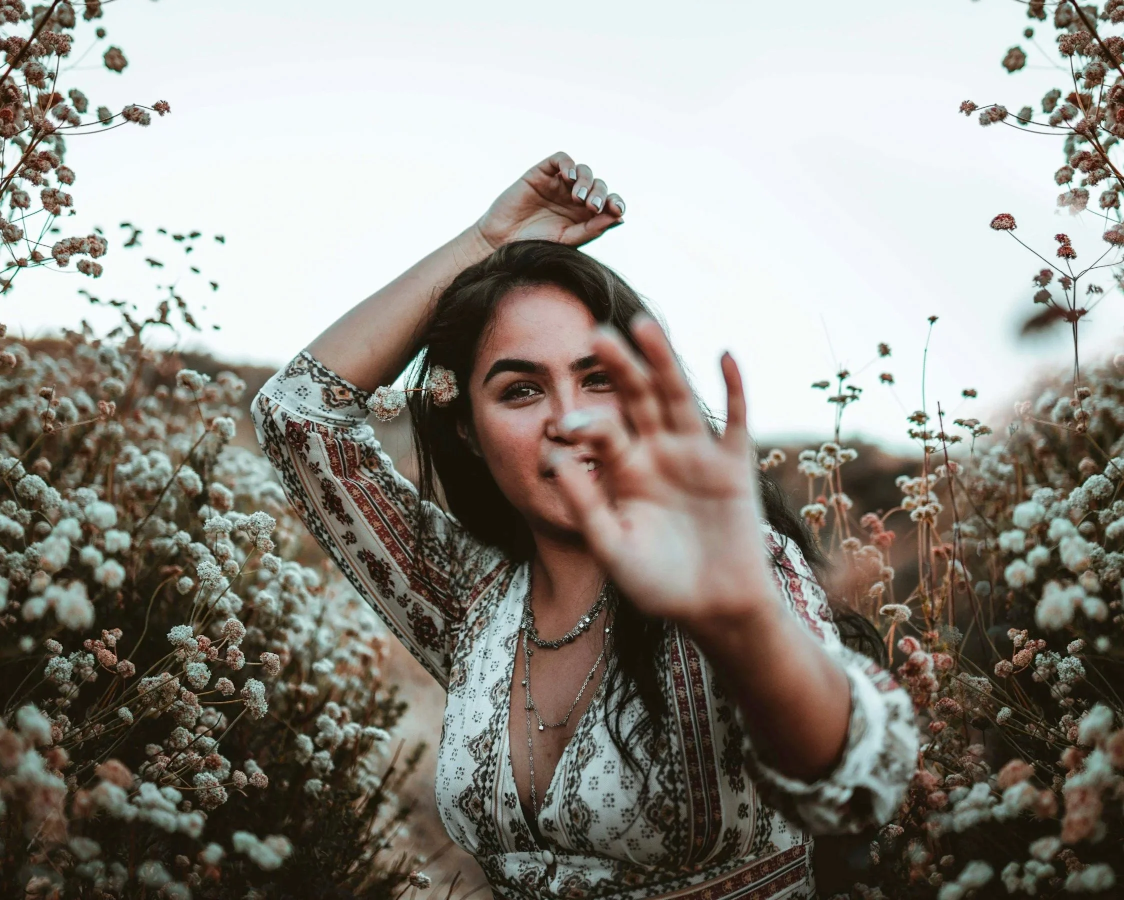 A woman with dark hair and patterned blouse standing in a field of small white flowers, with one hand behind her head and the other reaching out towards the camera.